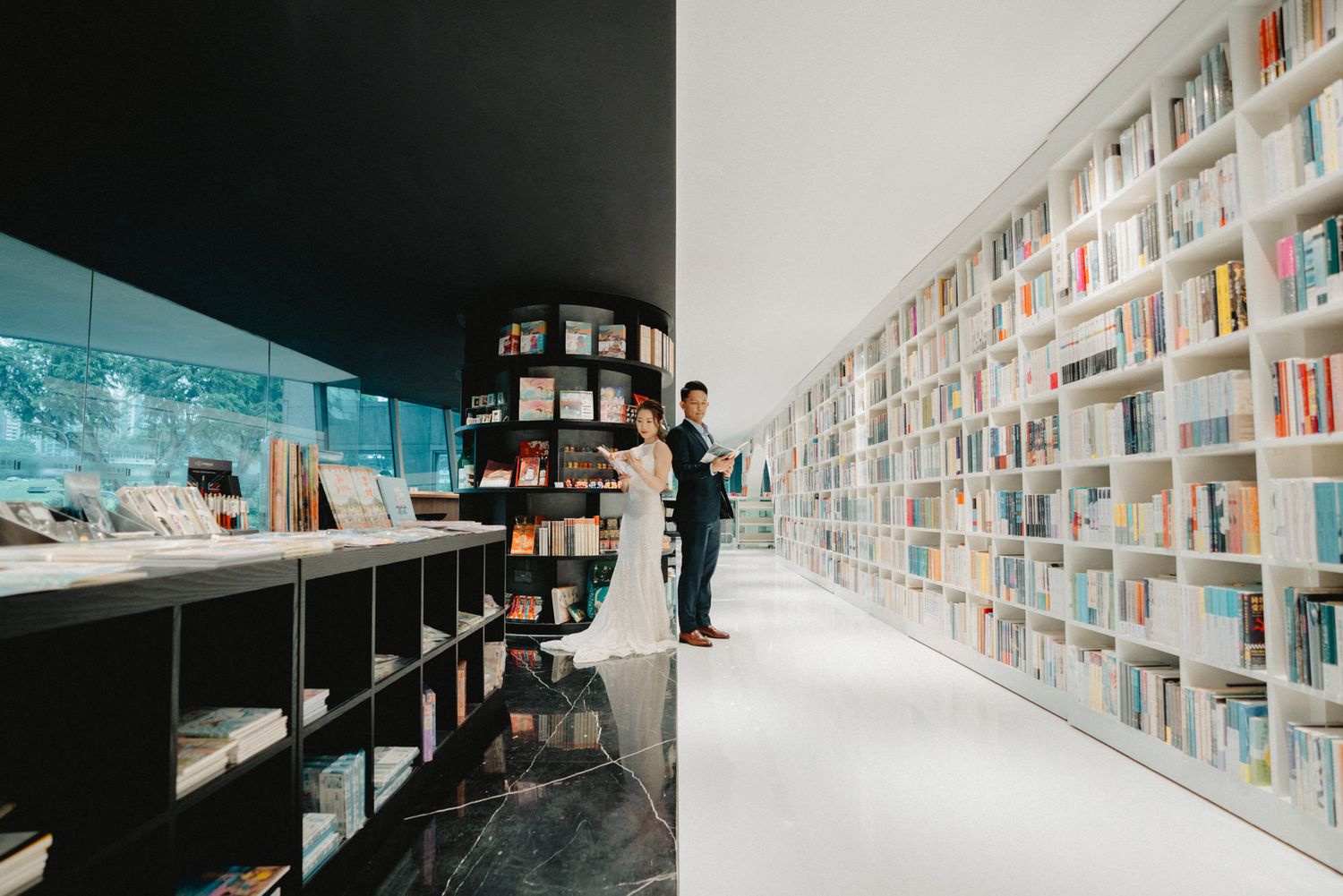 Couple poses in a modern bookstore with floor-to-ceiling white shelves and dark wood shelving during wedding photos.