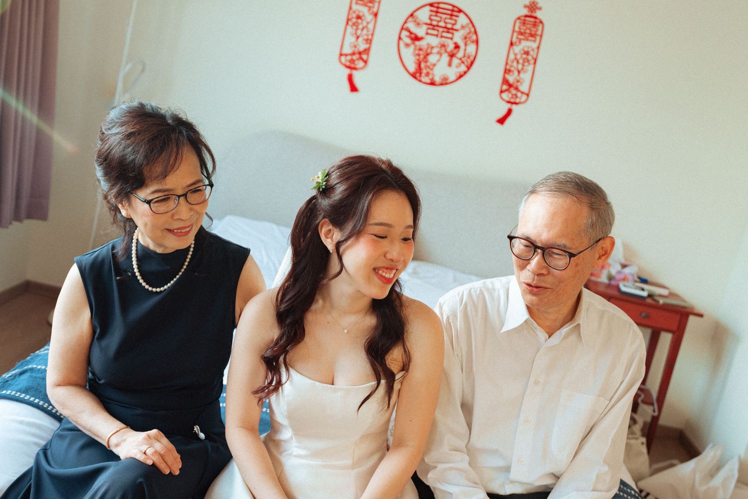 Bride in white dress smiles with elderly parents on bed, Chinese paper-cut decorations on wall behind them.