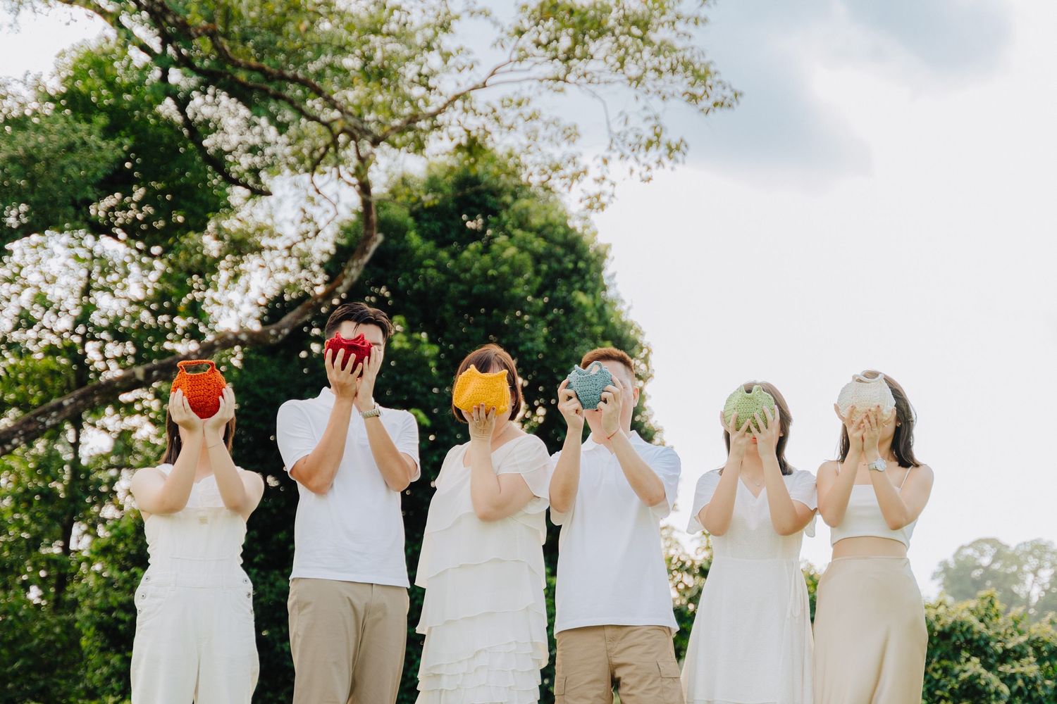 A group of people in white tops stand in a row with their hands raised to their faces in a synchronized pose outdoors.