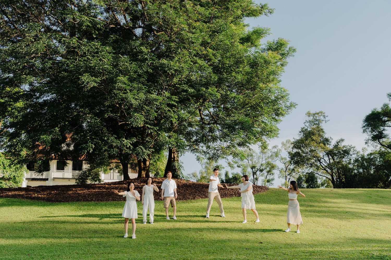 People in white outfits play croquet on a lush green lawn surrounded by trees on a sunny summer day.