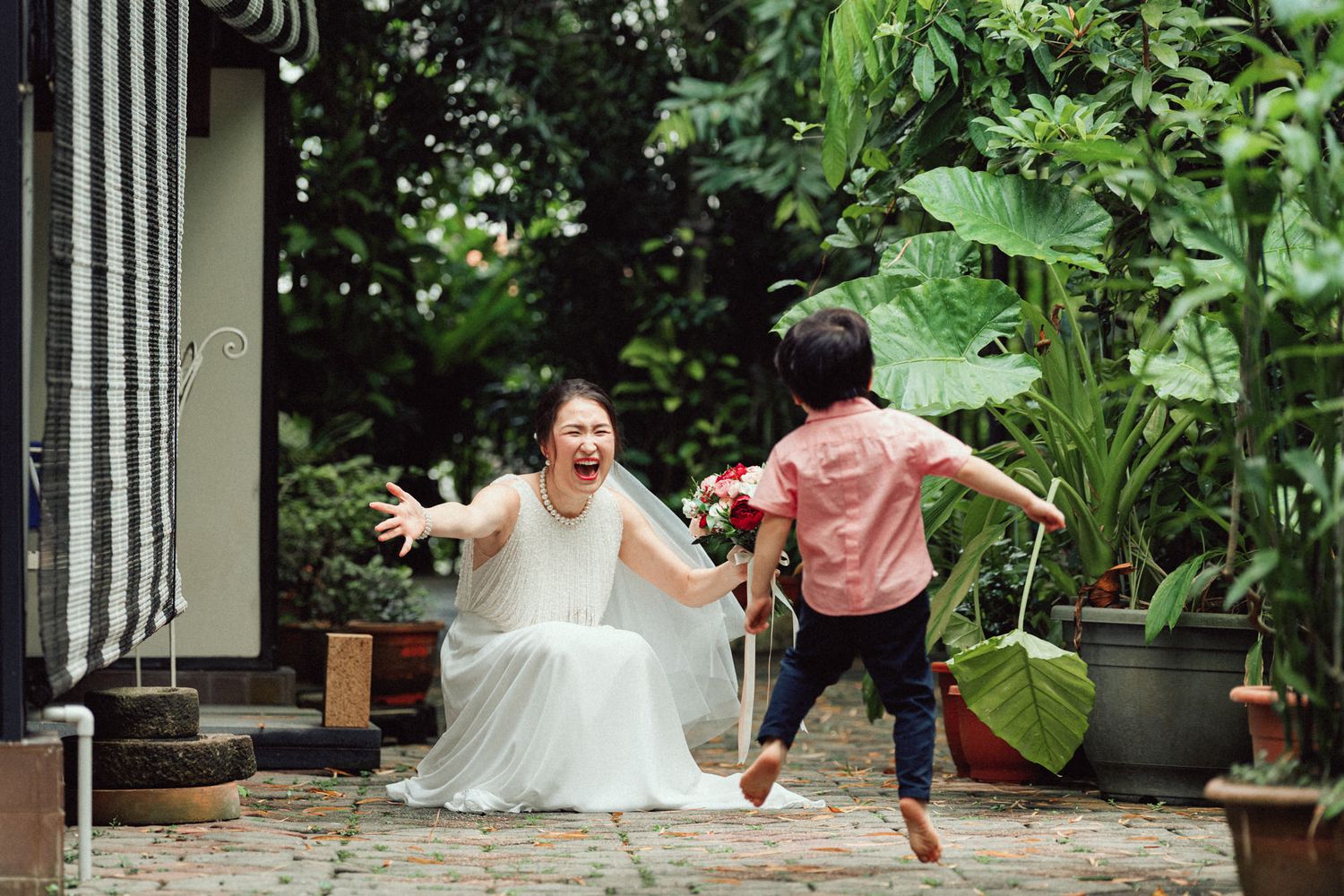 Joyful bride in white dress laughing as excited child jumps toward her in lush green garden setting.