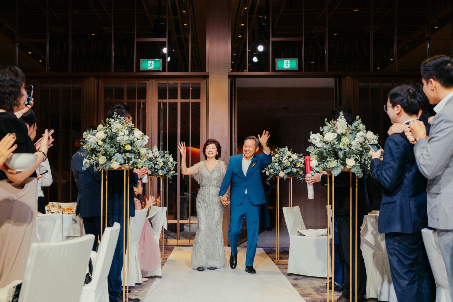 Couple walks down wedding aisle waving to guests, flanked by tall gold floral stands with white blooms in elegant venue.
