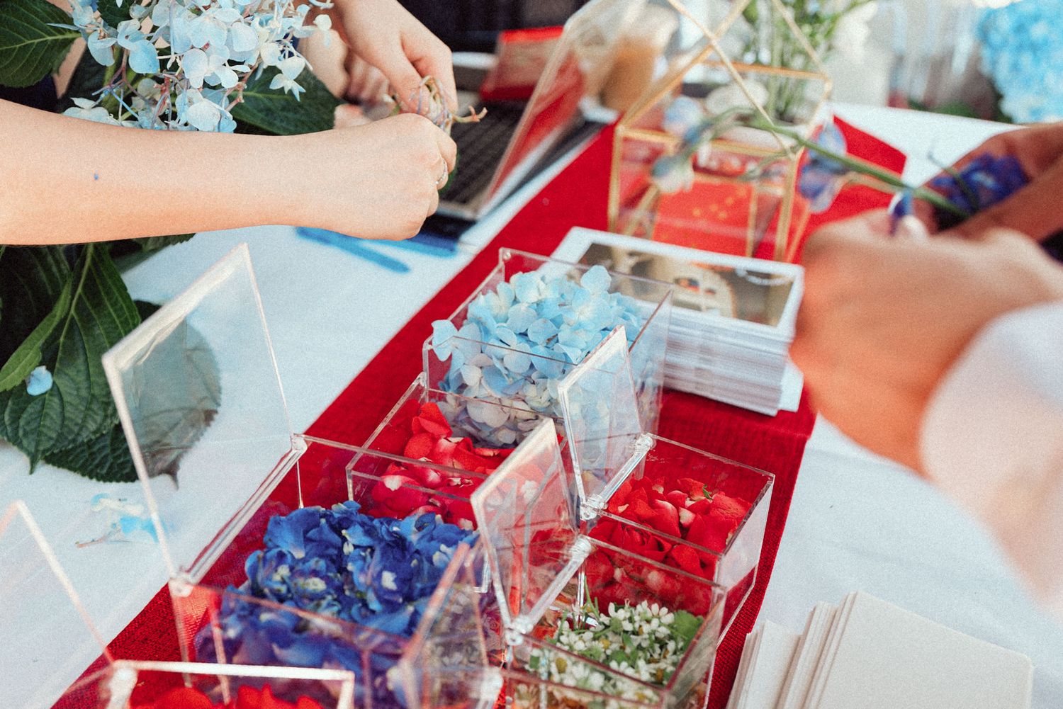 Hands arranging blue flowers and craft supplies in red and white compartmentalized organizer boxes.