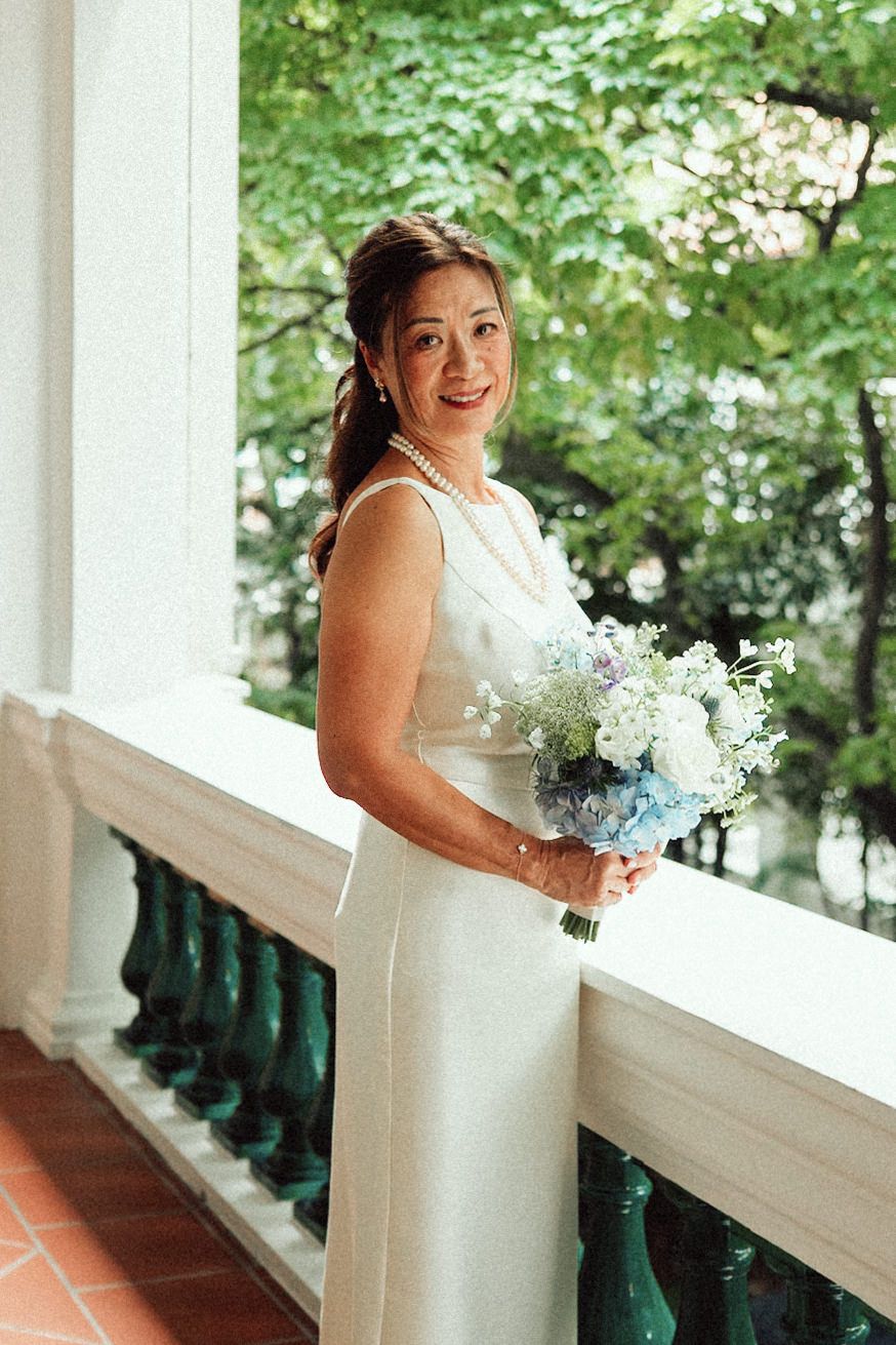 A bride in a sleek white wedding dress holds a blue and white bouquet while standing on a porch with green foliage behind.