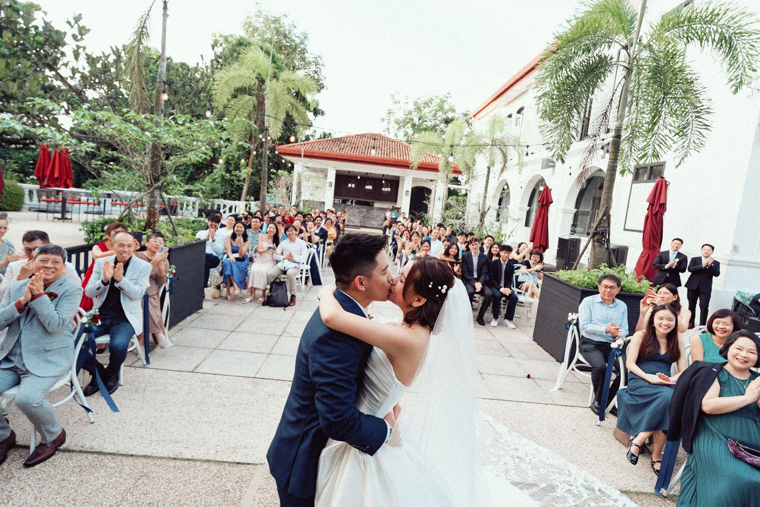 Wedding guests dancing together in an outdoor venue with palm trees and white buildings in the background.