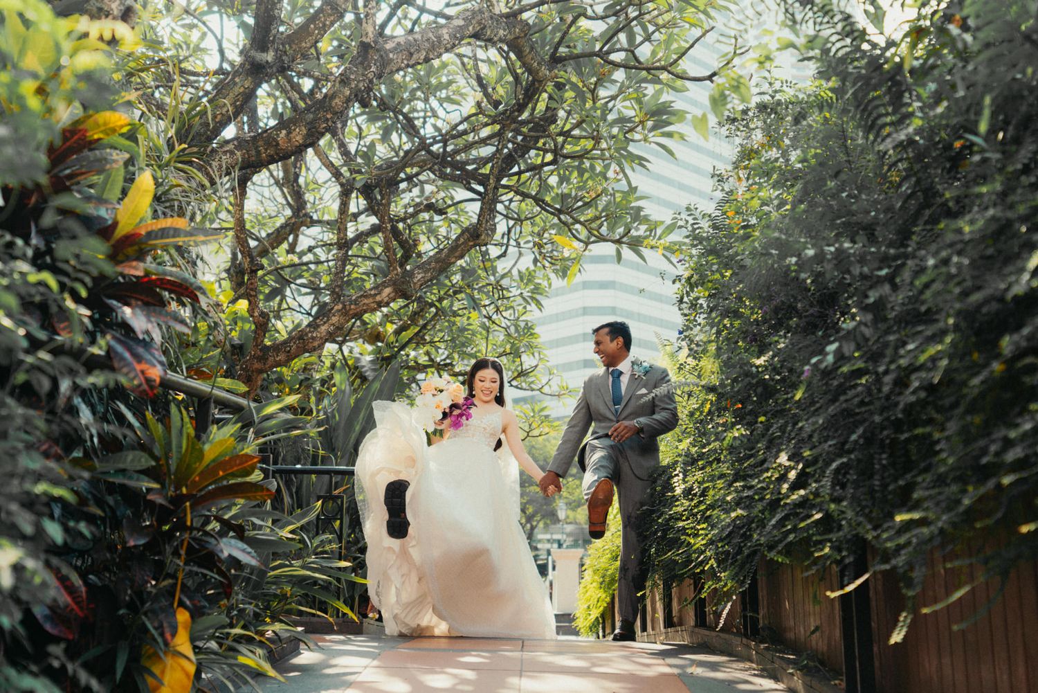 A bride and groom hold hands while walking through a lush green garden pathway on their wedding day.