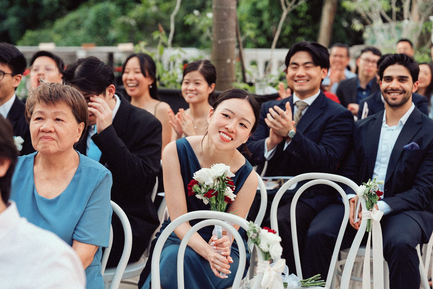 Wedding guests smile and laugh while seated in white chairs during an outdoor ceremony.