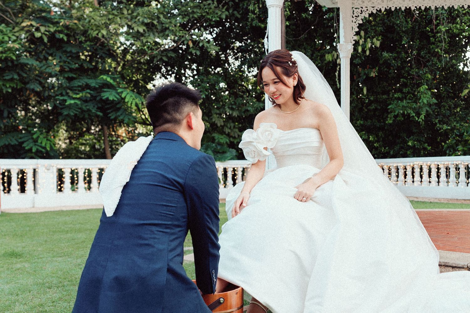 A couple in wedding attire shares a romantic moment on a garden lawn surrounded by greenery.