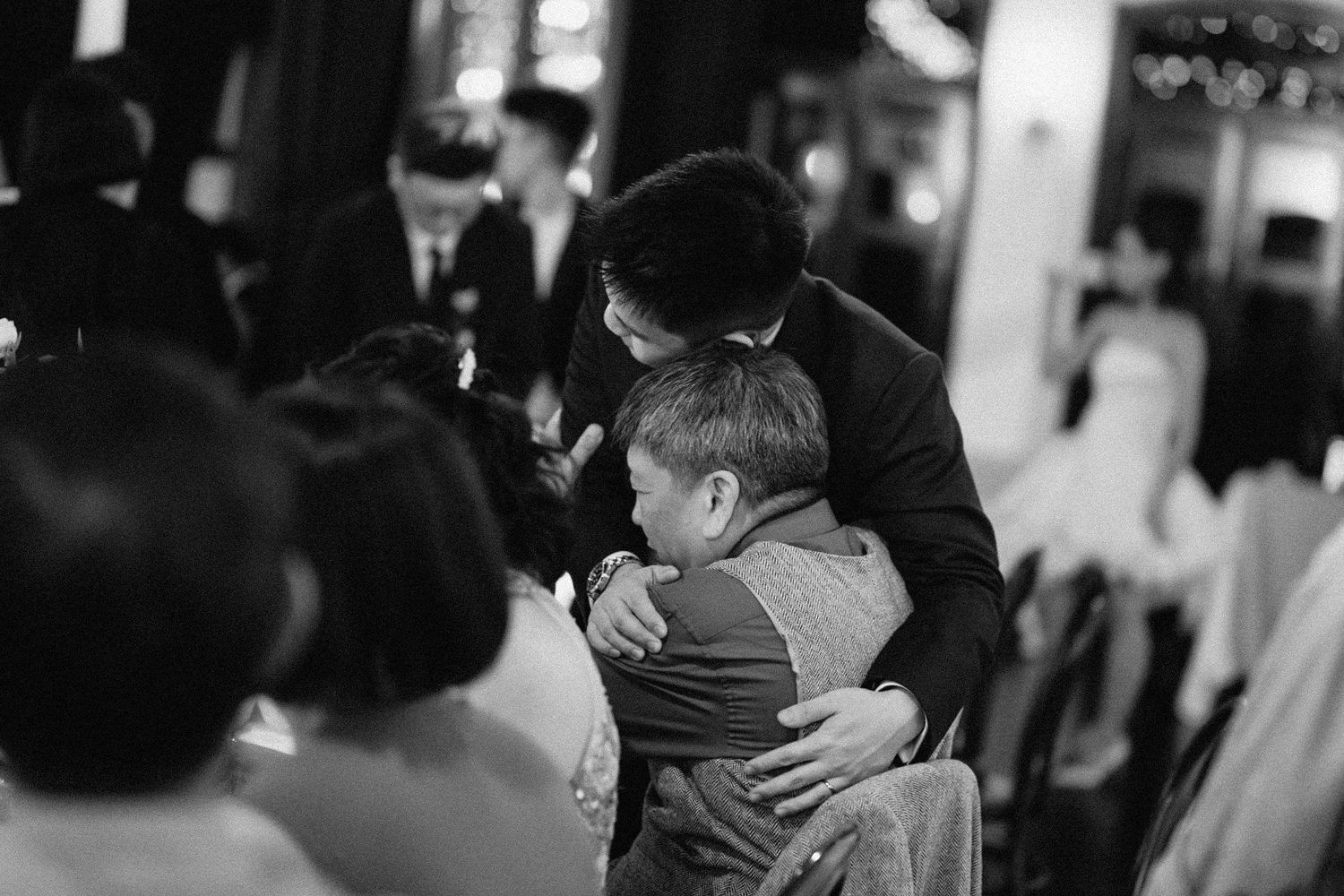 A tender black and white moment captured of a parent and child embracing in a crowded indoor setting.