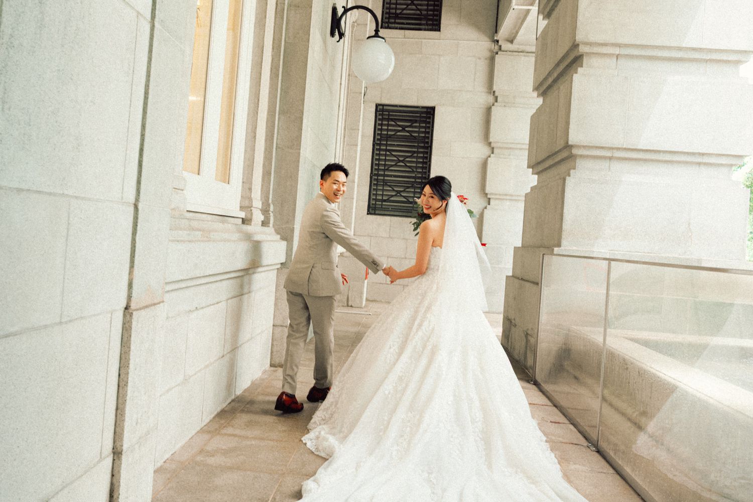 A bride in a flowing white gown and veil poses elegantly in a bright marble hallway with classical columns.