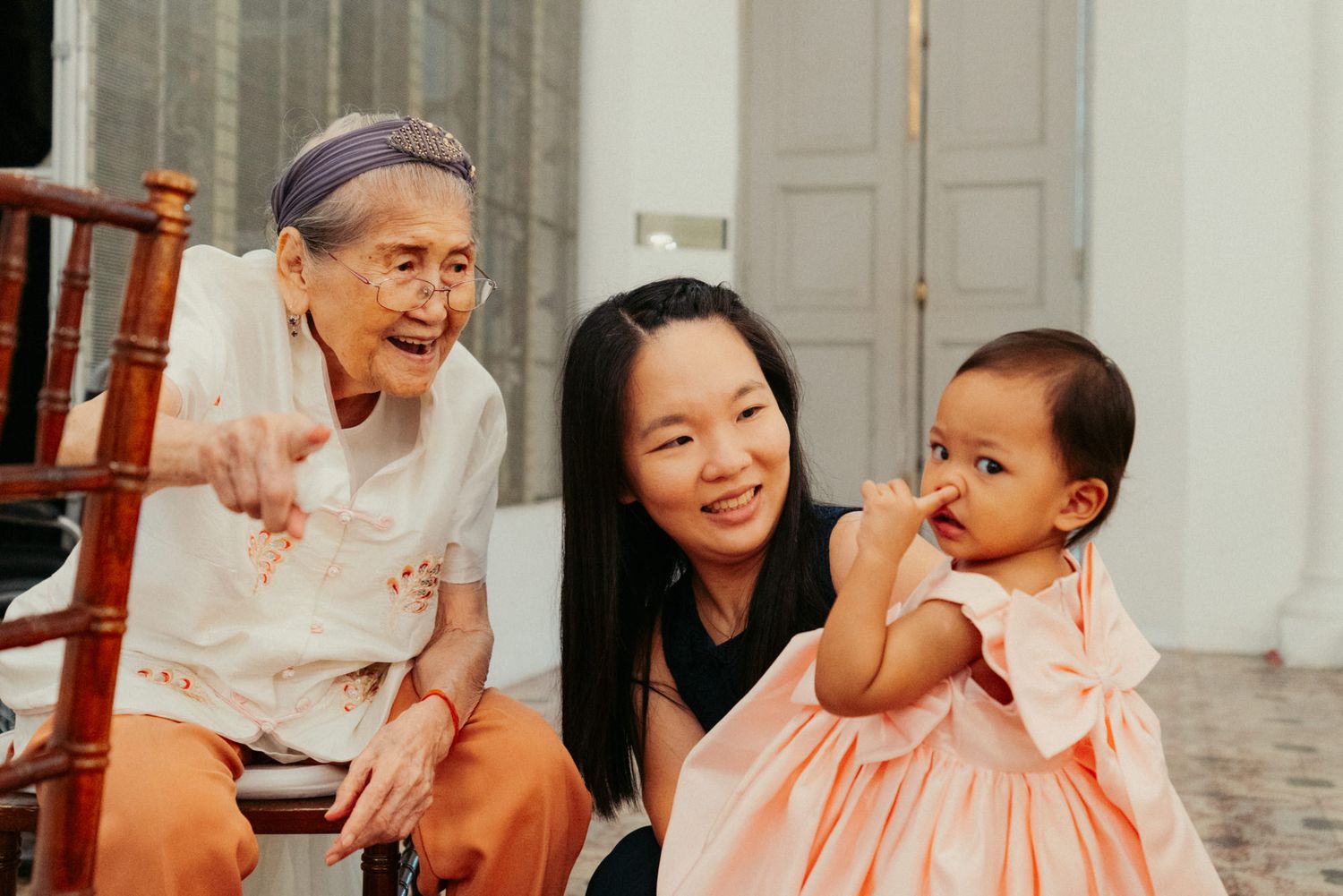 Three generations of women share a joyful moment, with an elderly woman, young mother, and baby girl in pink dress.