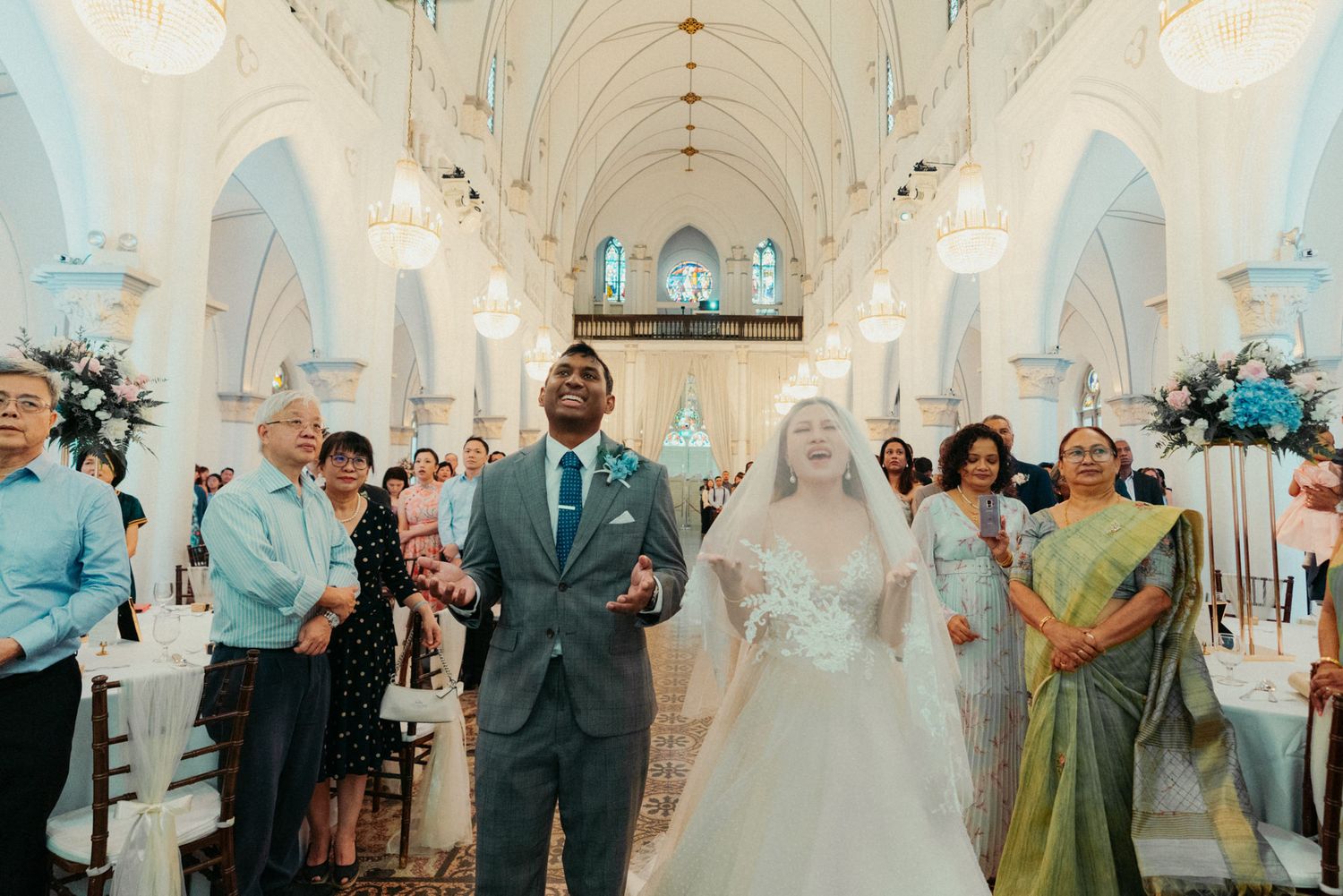 Groom and bride walk down the aisle of a stunning white church decorated with blue floral arrangements.