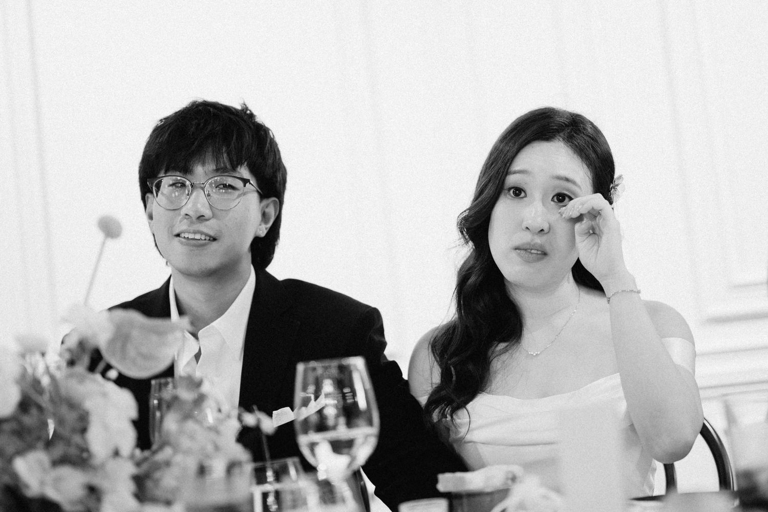 Black and white photo of a couple at a wedding reception, man in suit and woman in white dress wiping a tear.