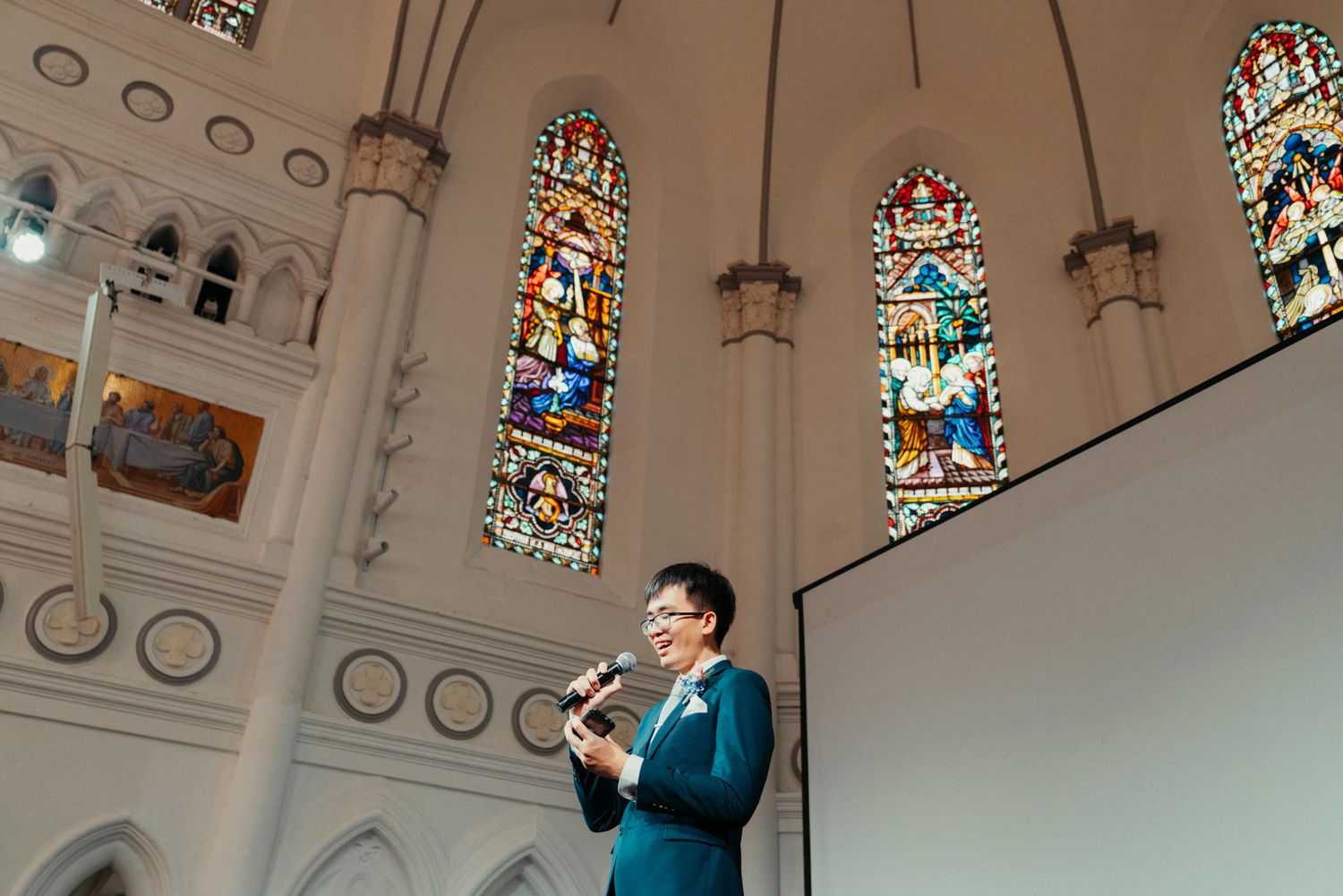 Woman speaks at podium inside elegant church with tall stained glass windows during wedding ceremony.