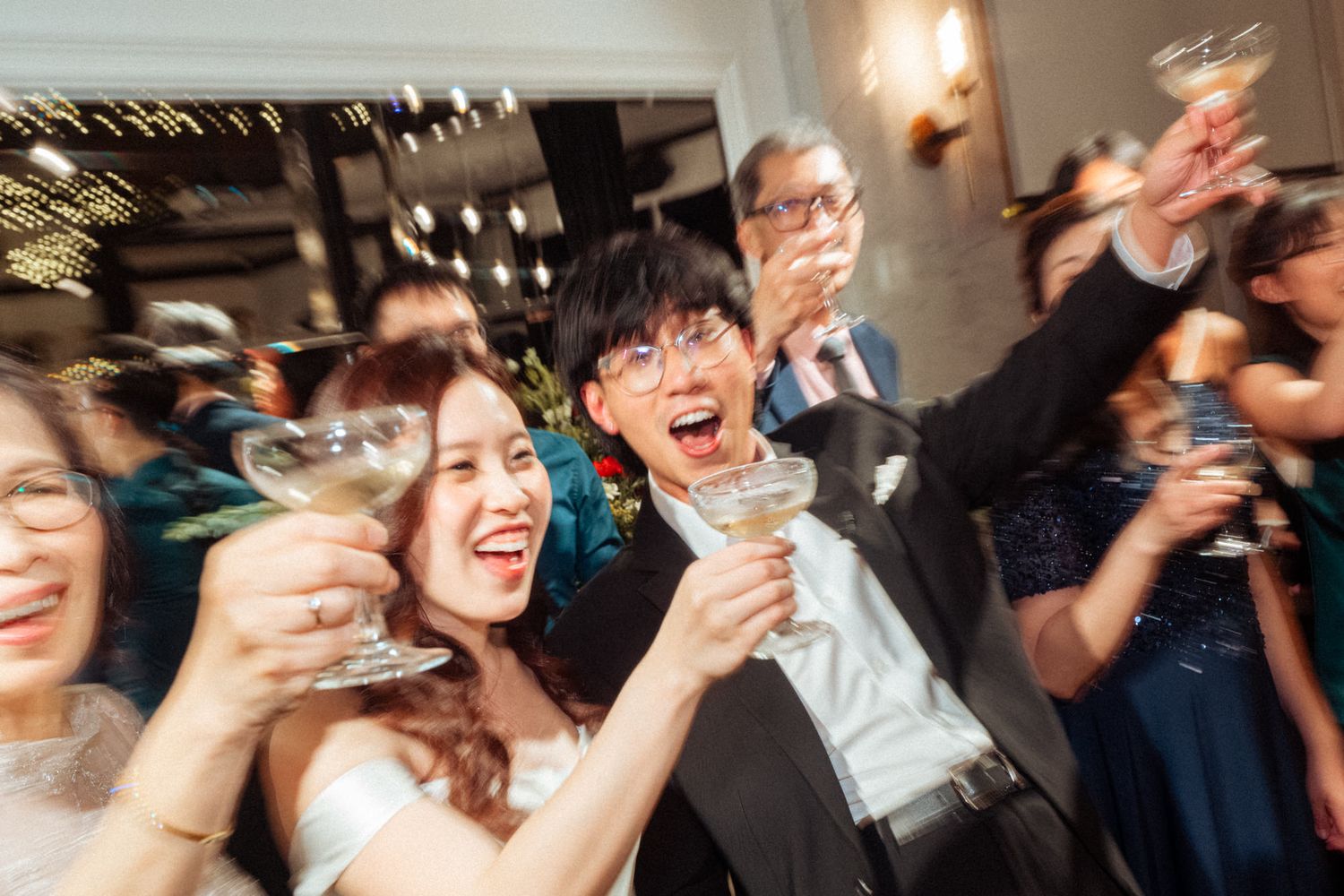Joyful wedding guests raising champagne glasses in a celebratory toast at a lively wedding reception.