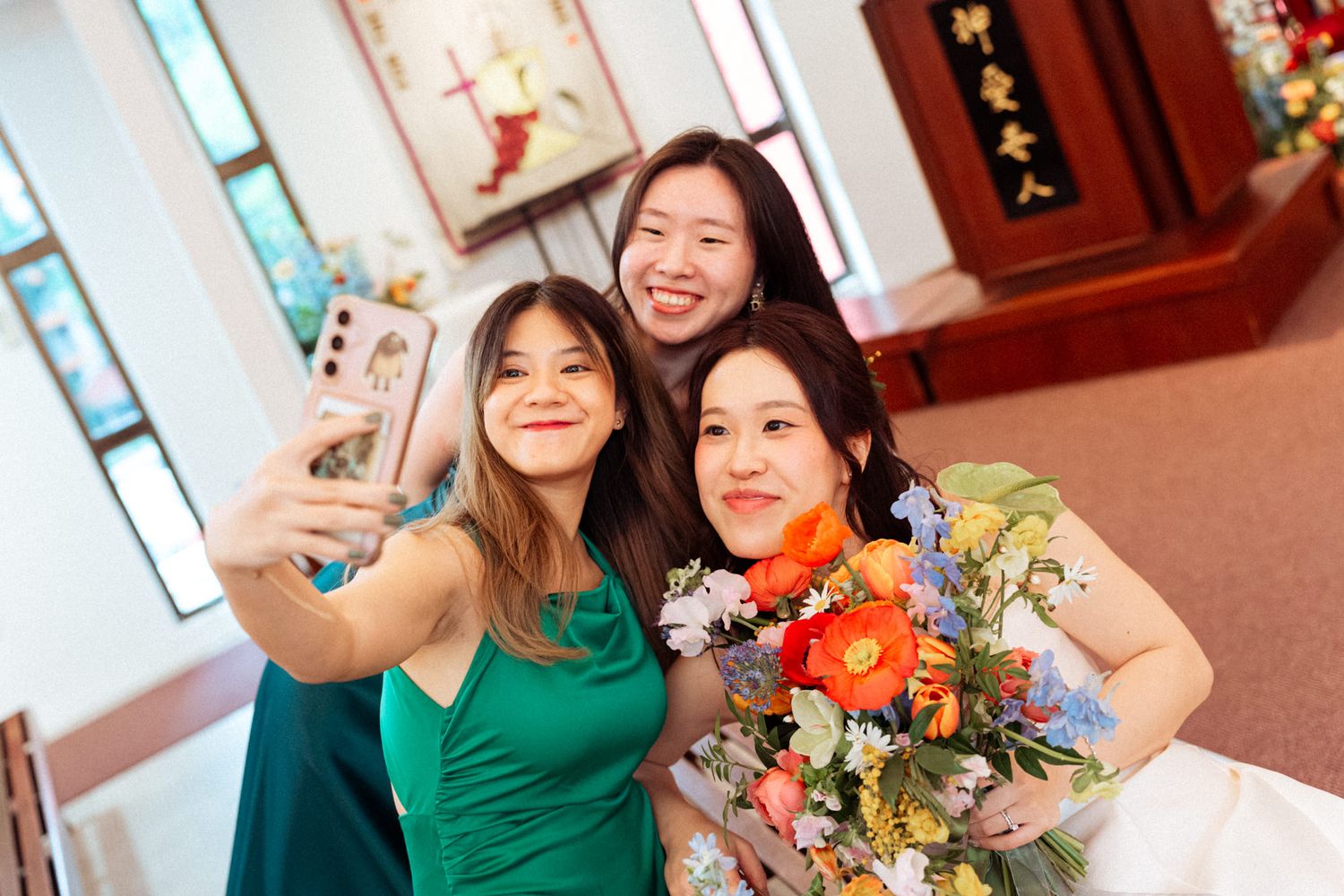 Three Asian women taking a selfie in a church, one holding a colorful bouquet of flowers.