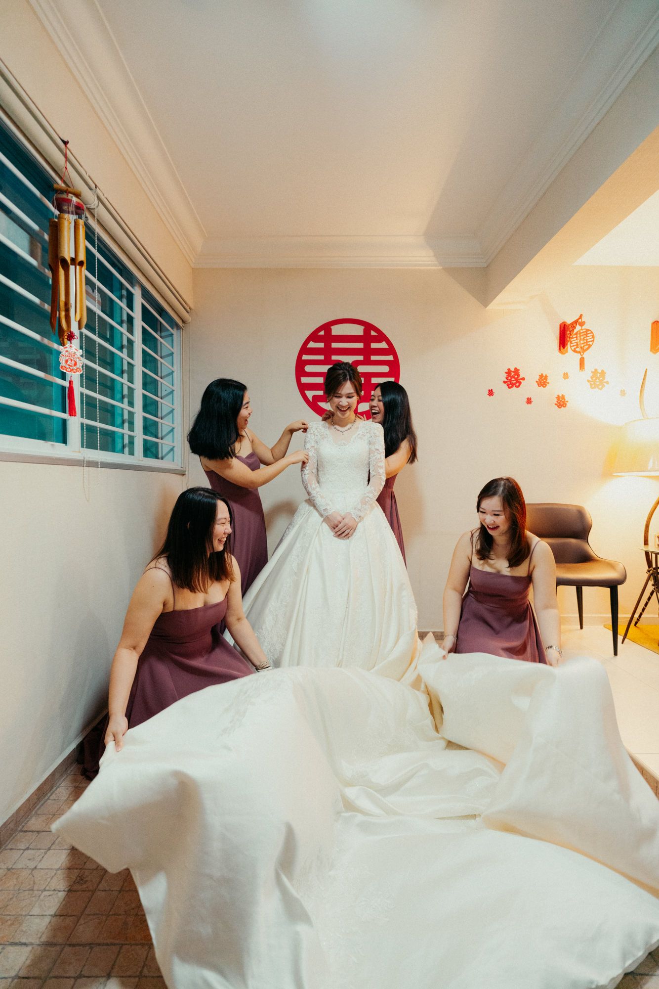 Bridesmaids in burgundy dresses help bride in white gown prepare for Chinese wedding ceremony indoors.