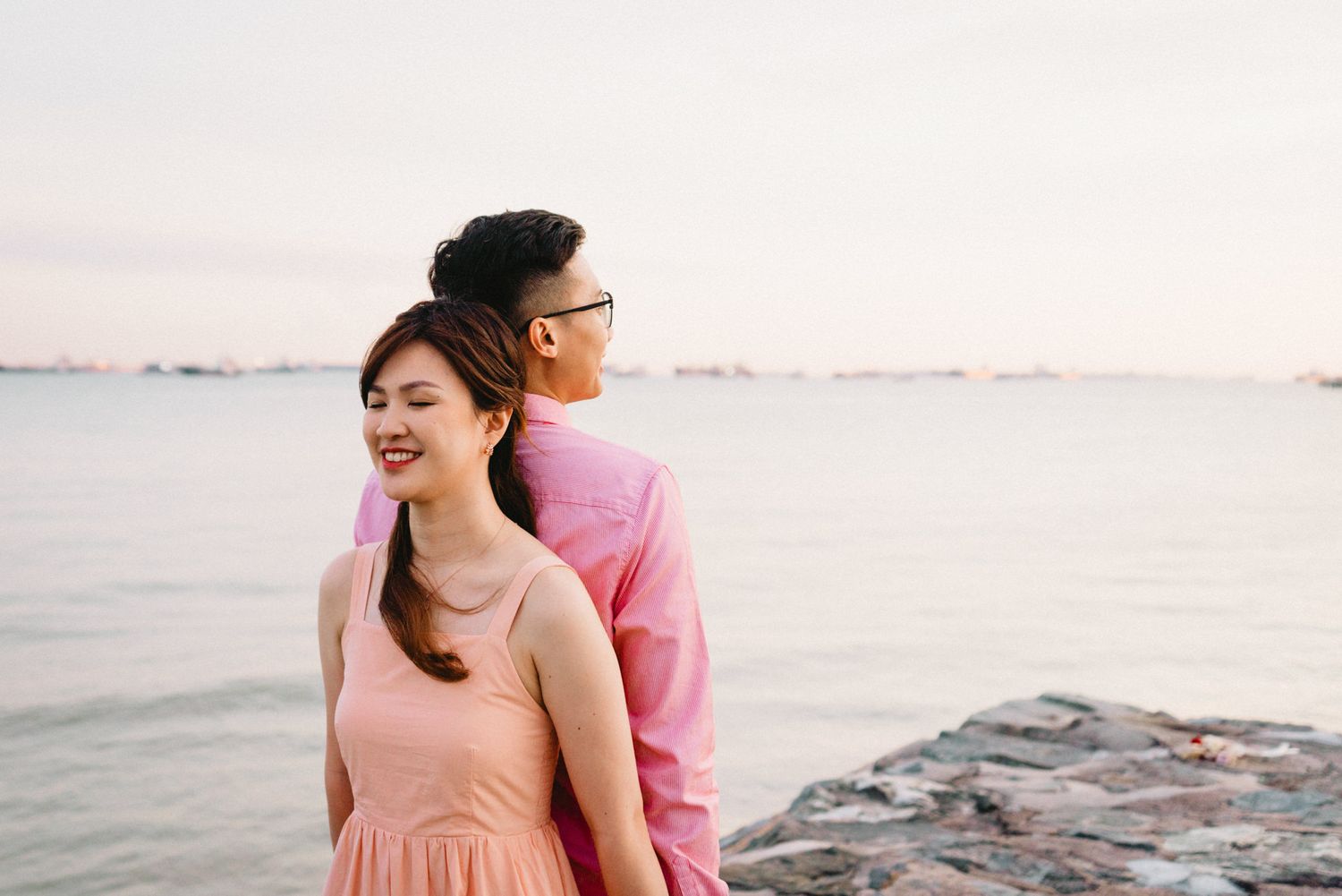 Couple in pink and peach outfits sharing romantic moments on a rocky beach during sunset.