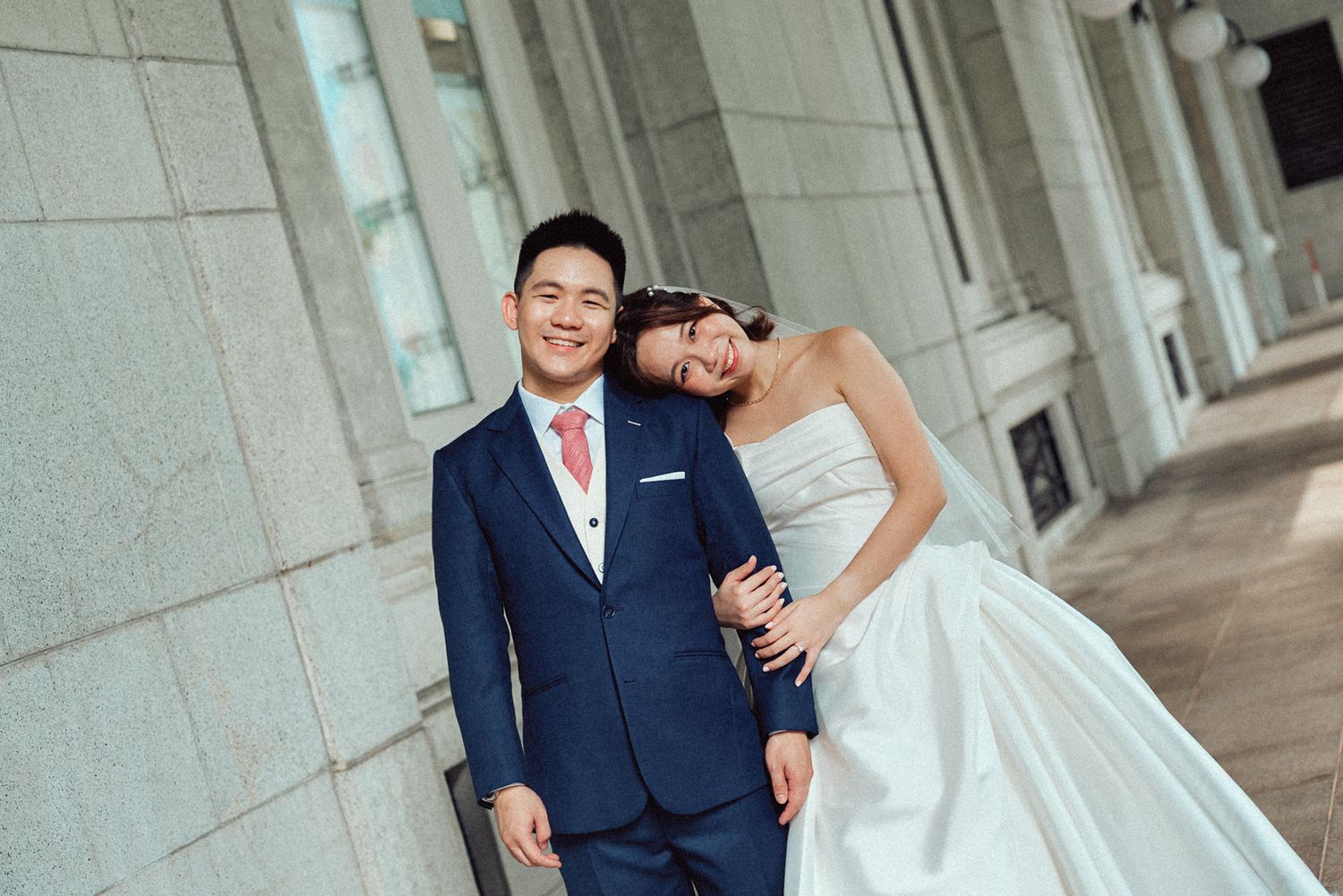 A couple in wedding attire poses together against a stone column exterior.