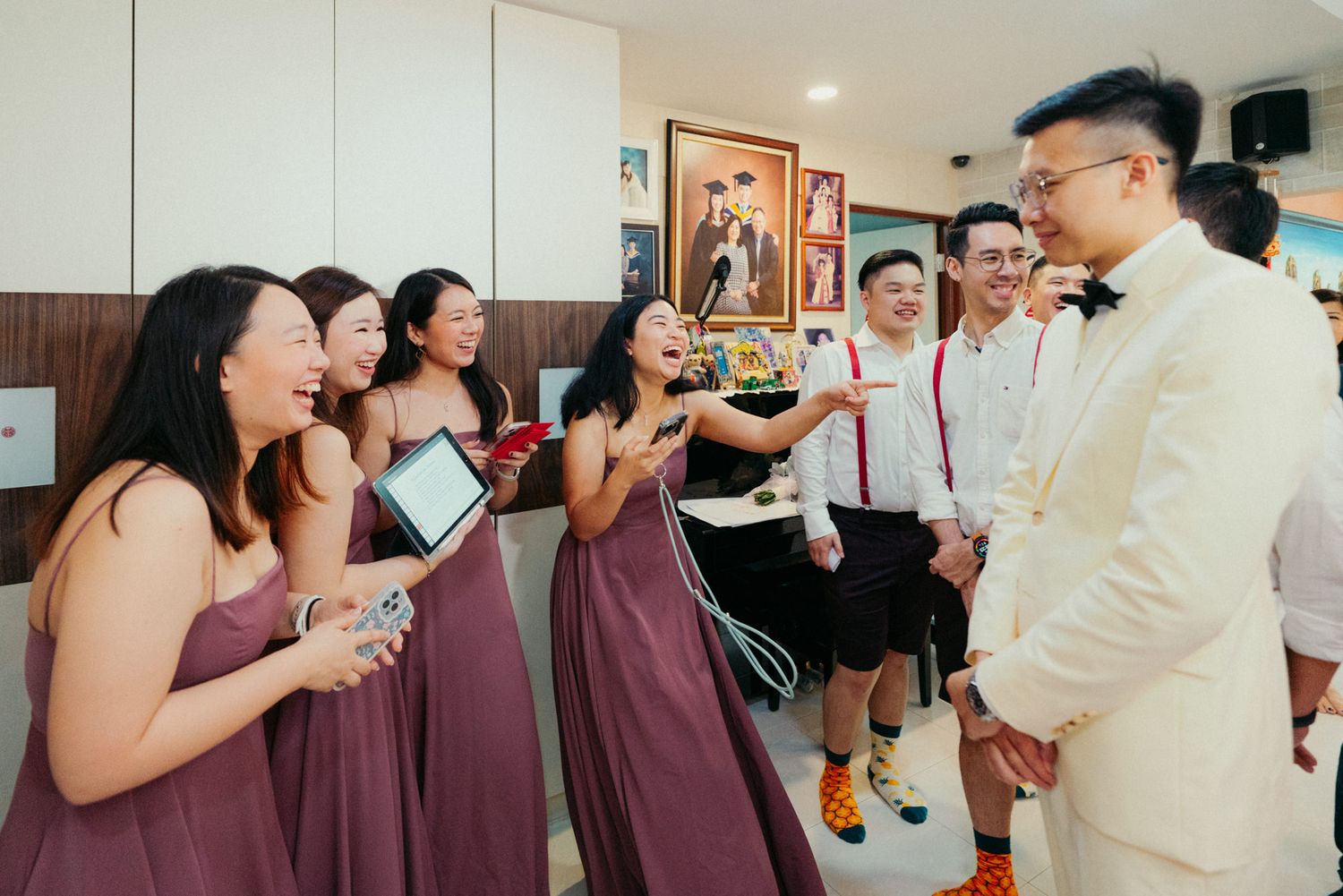 Bridesmaids in mauve dresses laugh with groomsmen in white shirts and red suspenders during a joyful wedding preparation moment.