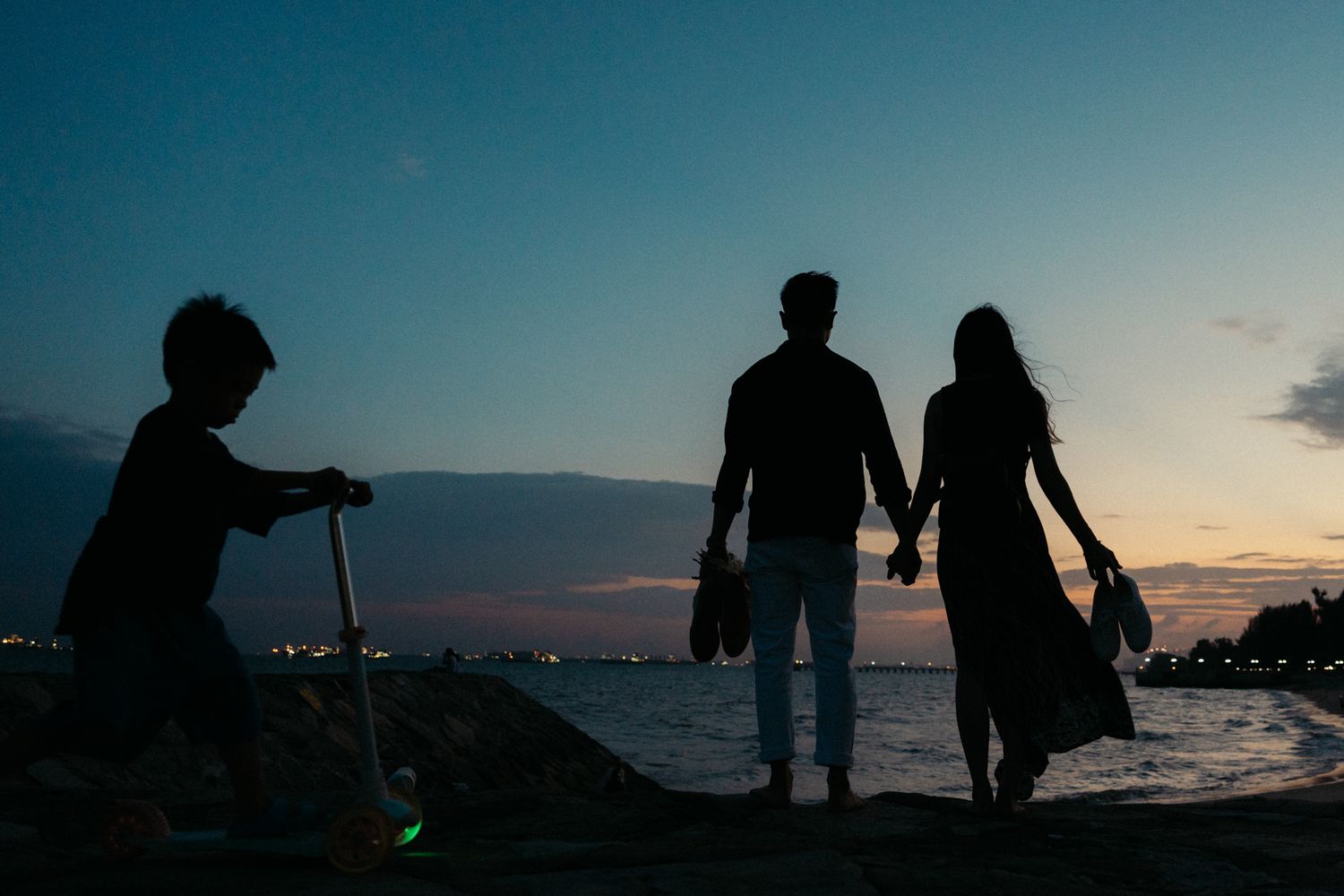 Silhouettes of couple walking on beach during sunset with city lights in background.