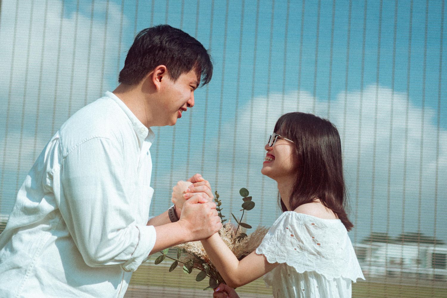 A couple in white outfits dances together in front of a blue sky with white clouds.