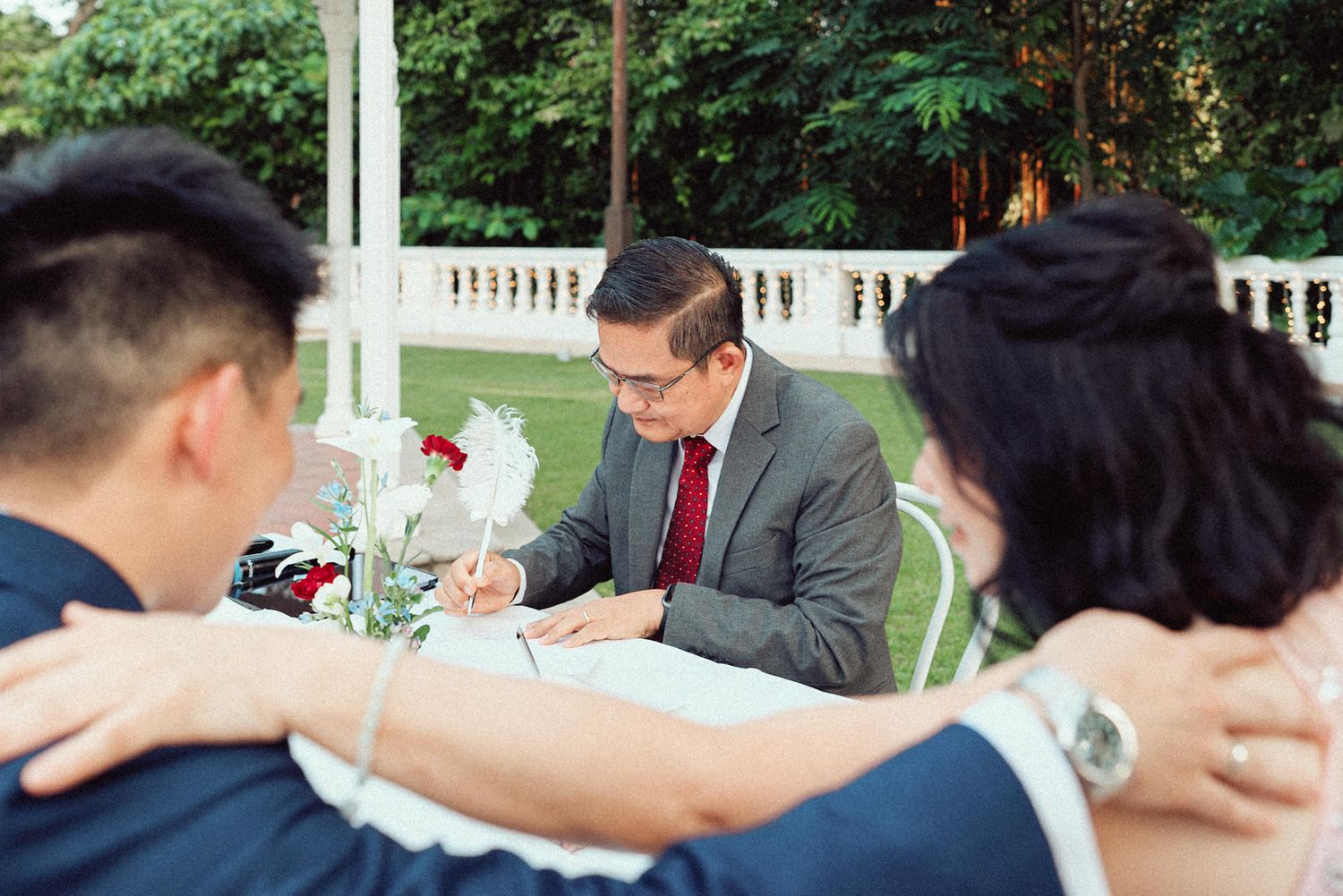 People sit at outdoor tables signing papers and documents during a gathering in a garden setting.