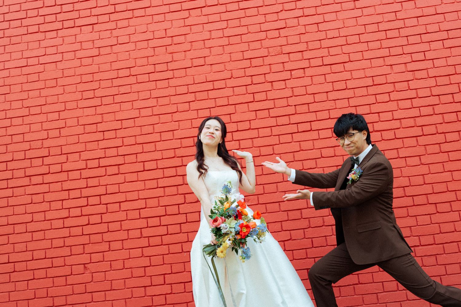 Bride in white gown holds colorful bouquet while groom in brown suit playfully poses against a red brick wall.