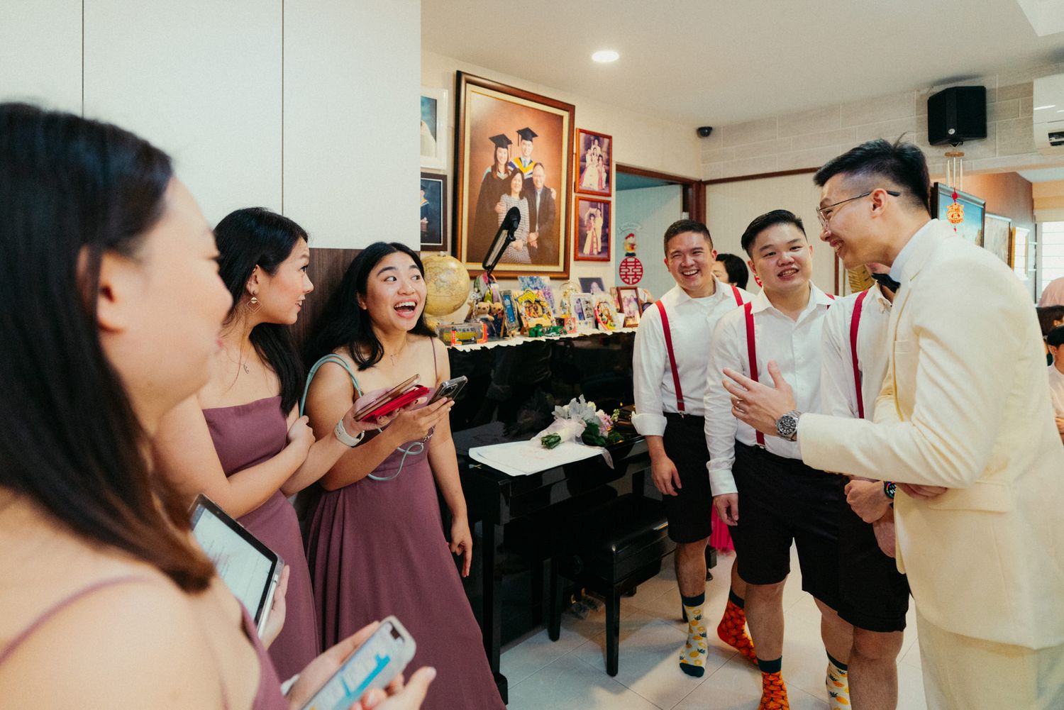 Wedding party laughing and celebrating indoors, bridesmaids in mauve dresses with groomsmen in white suits with red suspenders.