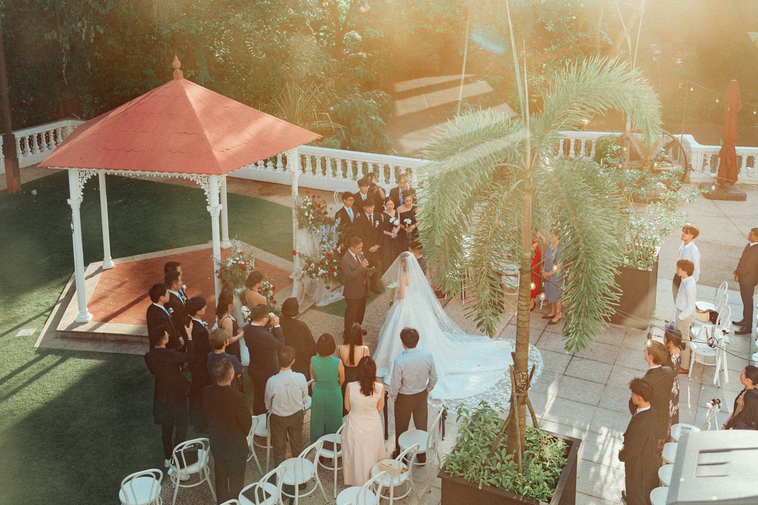 Aerial view of an elegant outdoor wedding ceremony near a red gazebo surrounded by palm trees and guests.