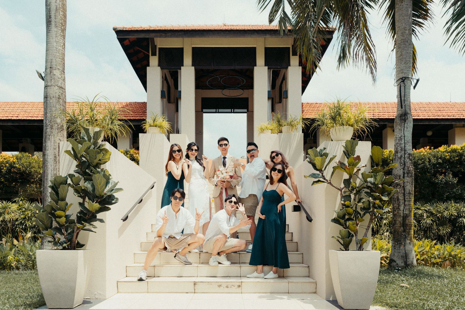 A wedding party poses on tropical resort steps, wearing elegant green and white attire with palm trees in background.