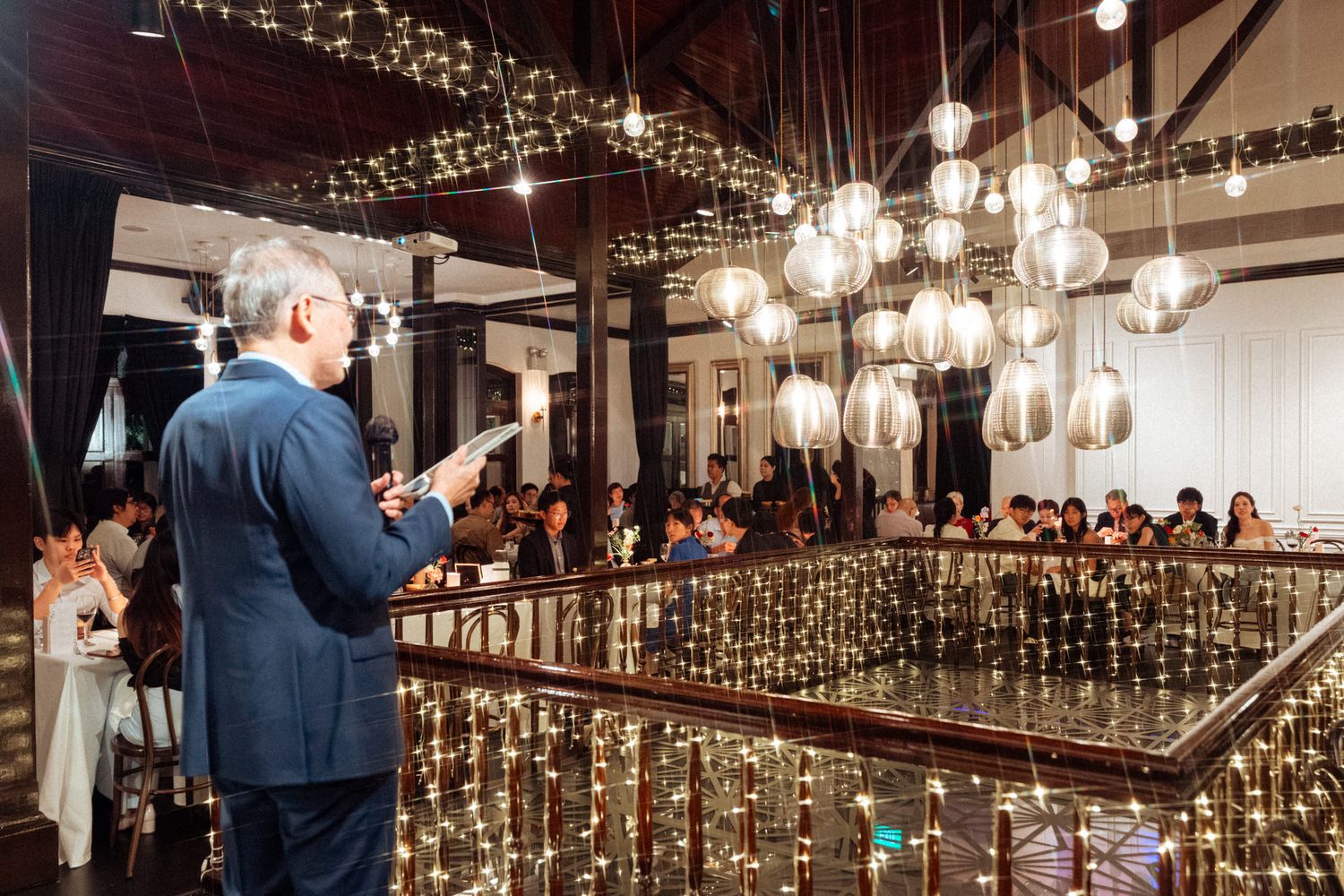 Man in blue suit speaks at elegant event venue with fairy lights and globe chandeliers to seated guests.