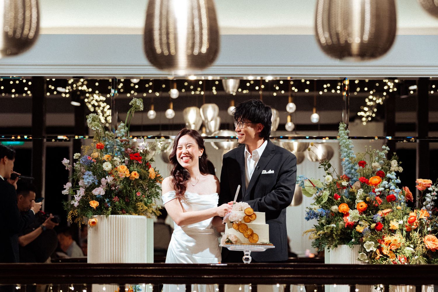 A joyful couple cuts their wedding cake surrounded by colorful floral arrangements and twinkling lights at an elegant venue.