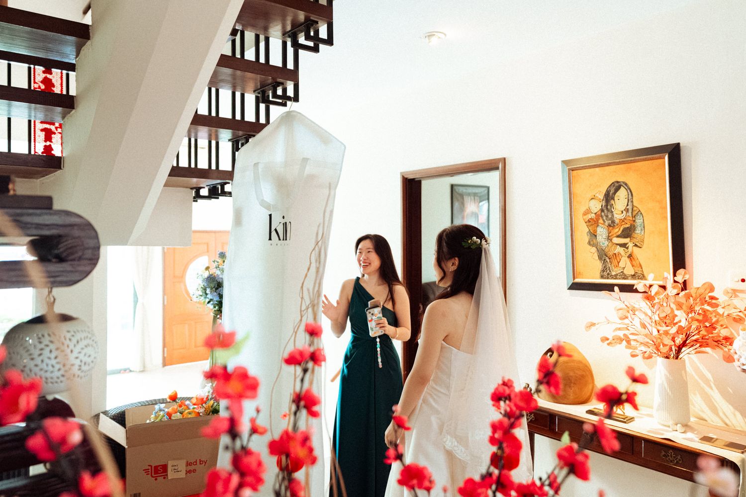 Two women in elegant dresses discuss bridal gown details in a beautifully decorated home with red floral accents.