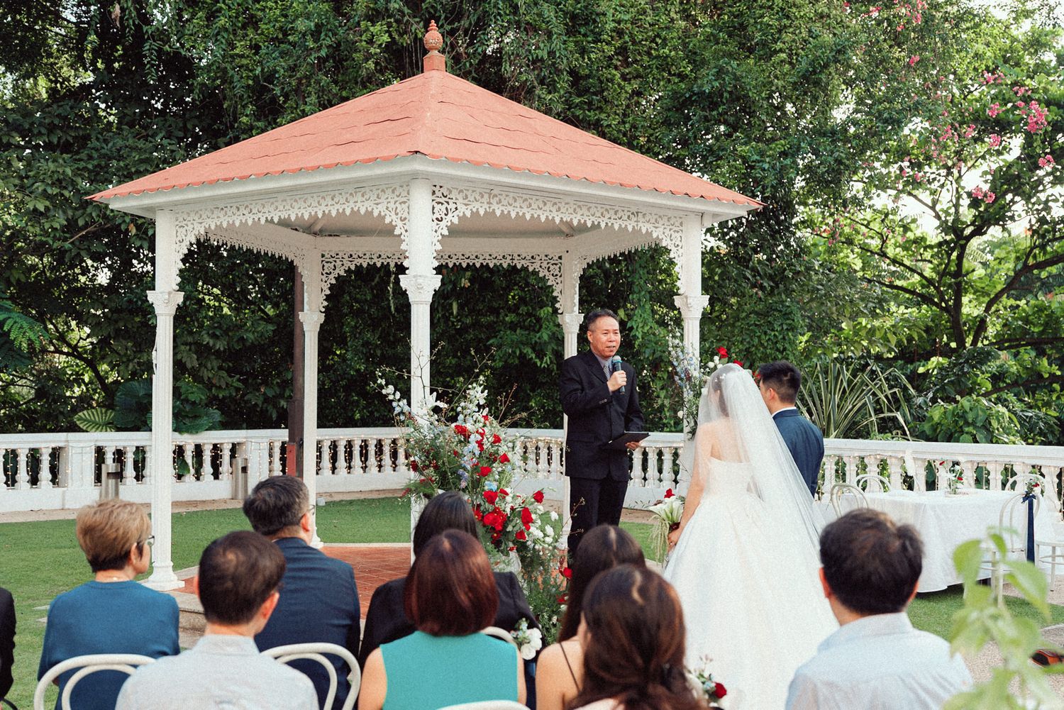 A wedding ceremony takes place at an elegant white gazebo surrounded by greenery and guests in attendance.