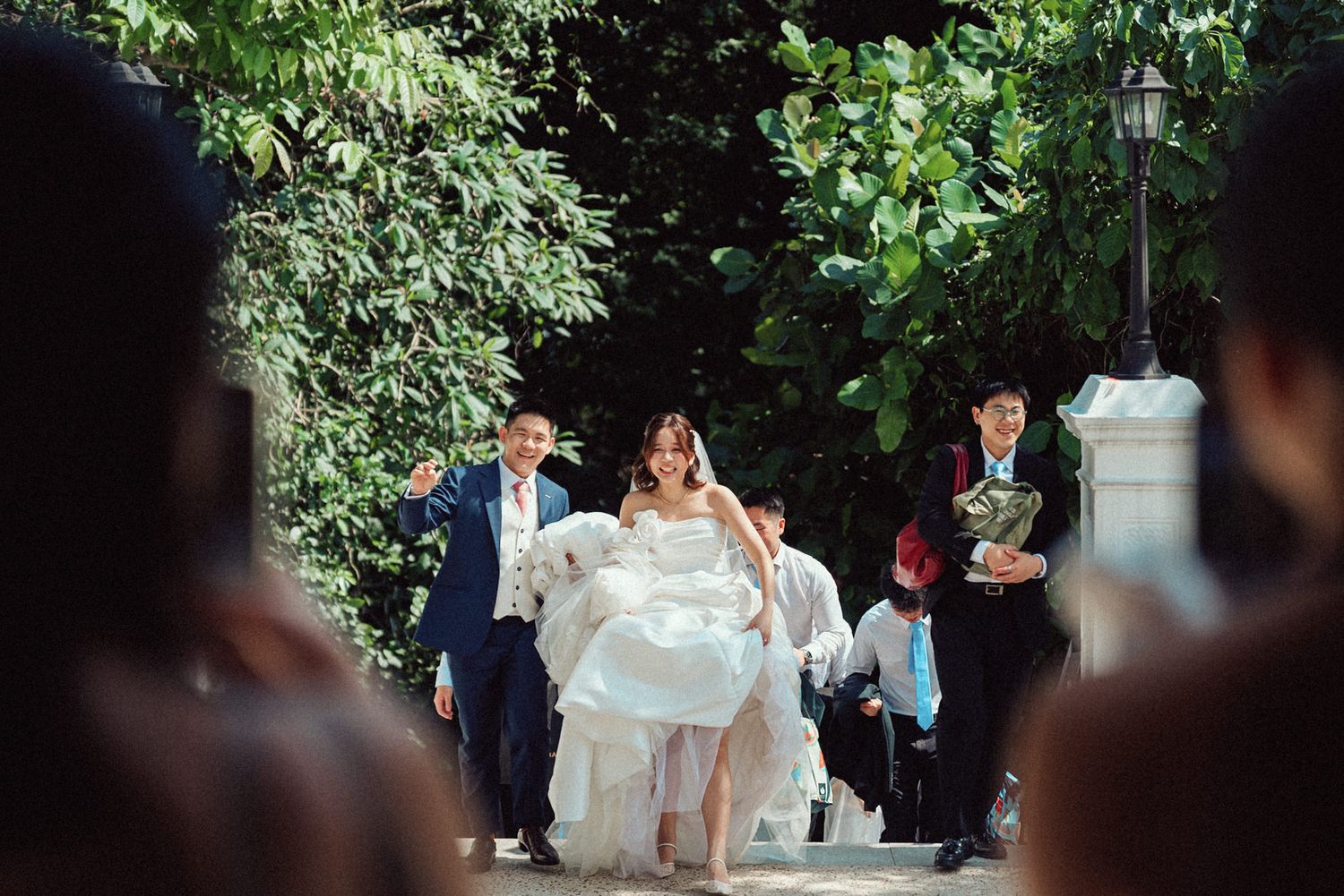 A couple walks through a lush garden pathway surrounded by green foliage during an outdoor event.