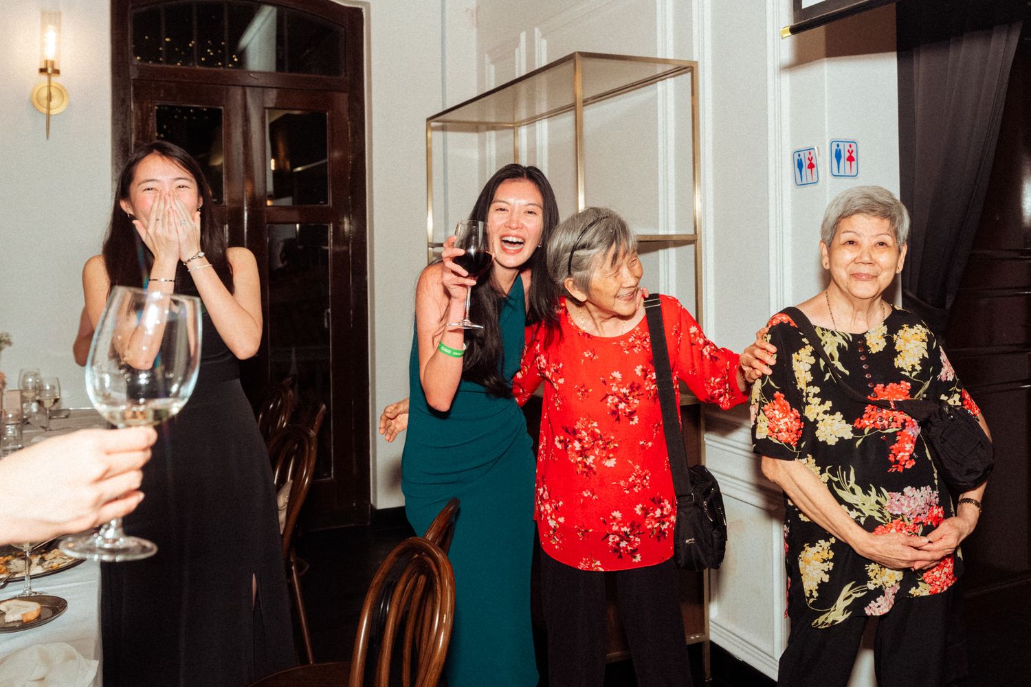 Four women laughing and celebrating at an elegant indoor party, holding wine glasses in a warmly lit dining room.