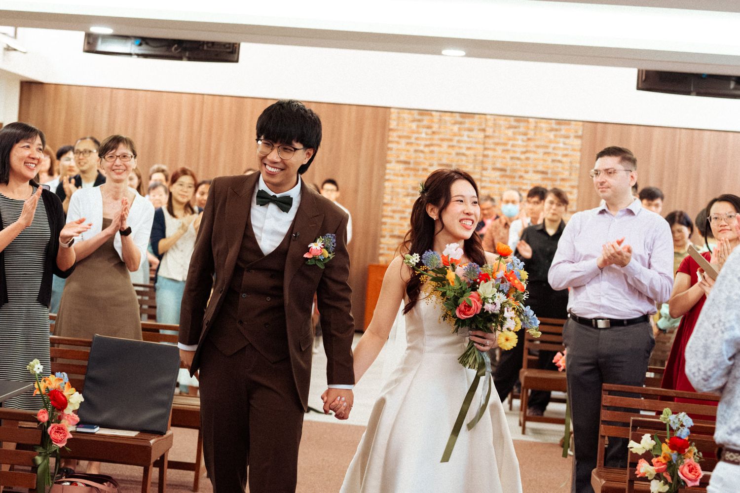A joyful couple walks down the aisle after their wedding ceremony in a chapel, greeted by applauding guests.