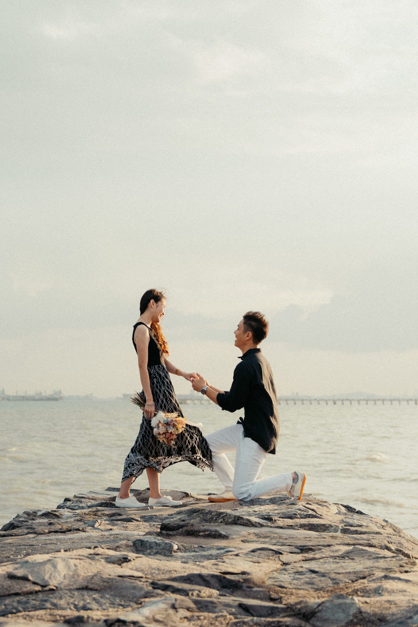 A couple creates romantic poses on rocky shoreline during sunset.