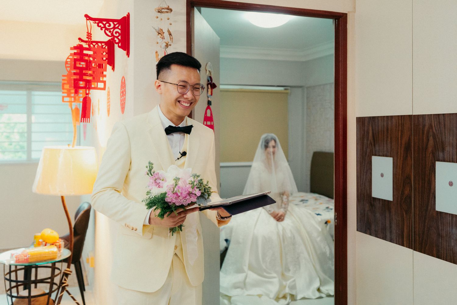 Groom in white suit holding flowers smiles as bride in wedding dress waits in background, Chinese wedding decorations visible.