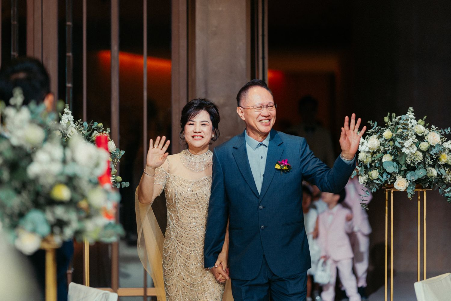 A smiling couple in formal attire waves at a wedding reception entrance, surrounded by floral arrangements.