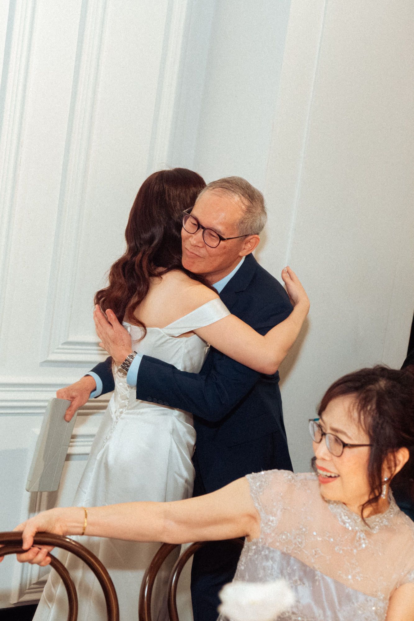 A bride in a white gown shares a heartfelt hug with an older man in a suit at a wedding reception.
