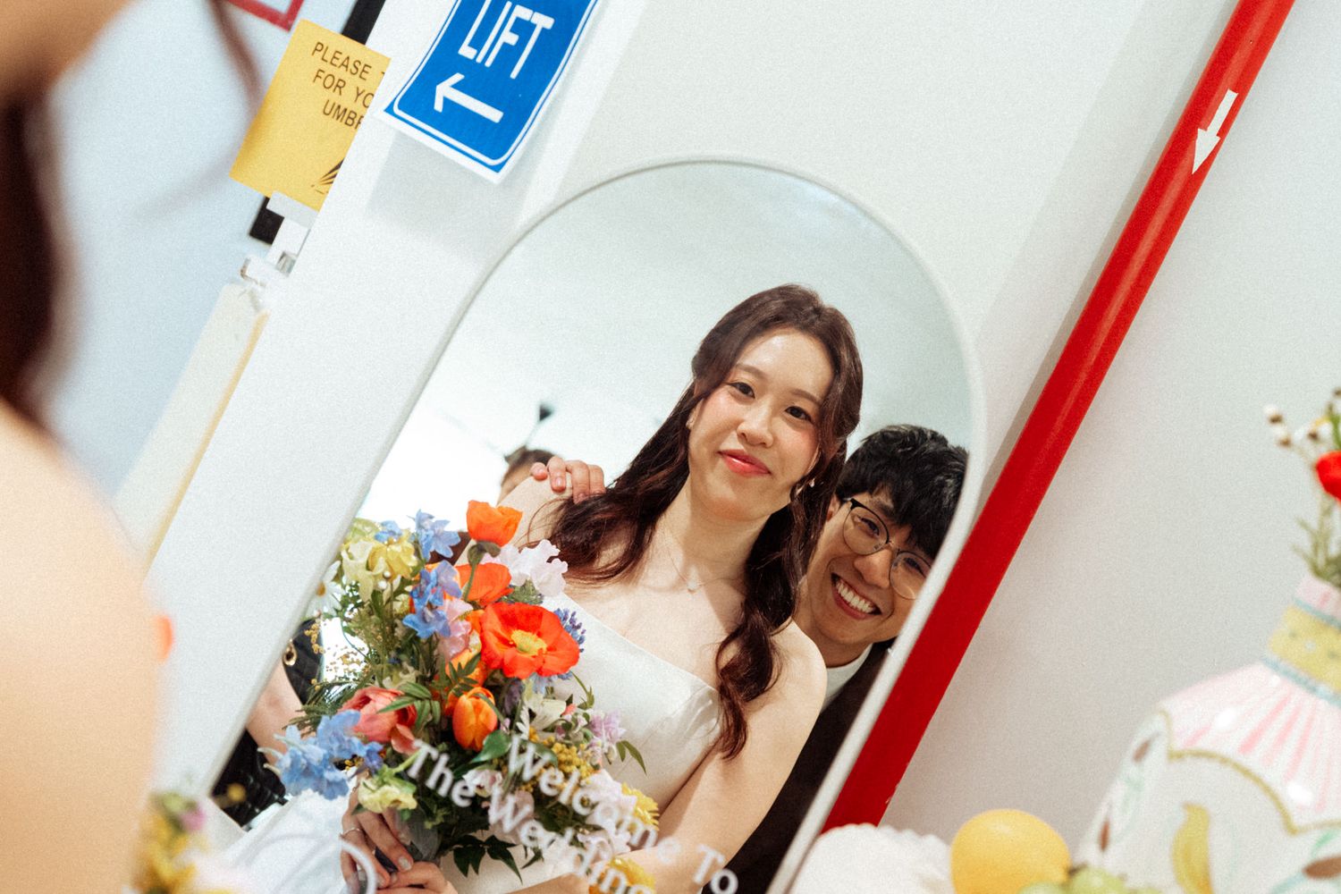 Bride in white dress holding colorful bouquet poses with guest reflected in oval mirror at wedding venue.
