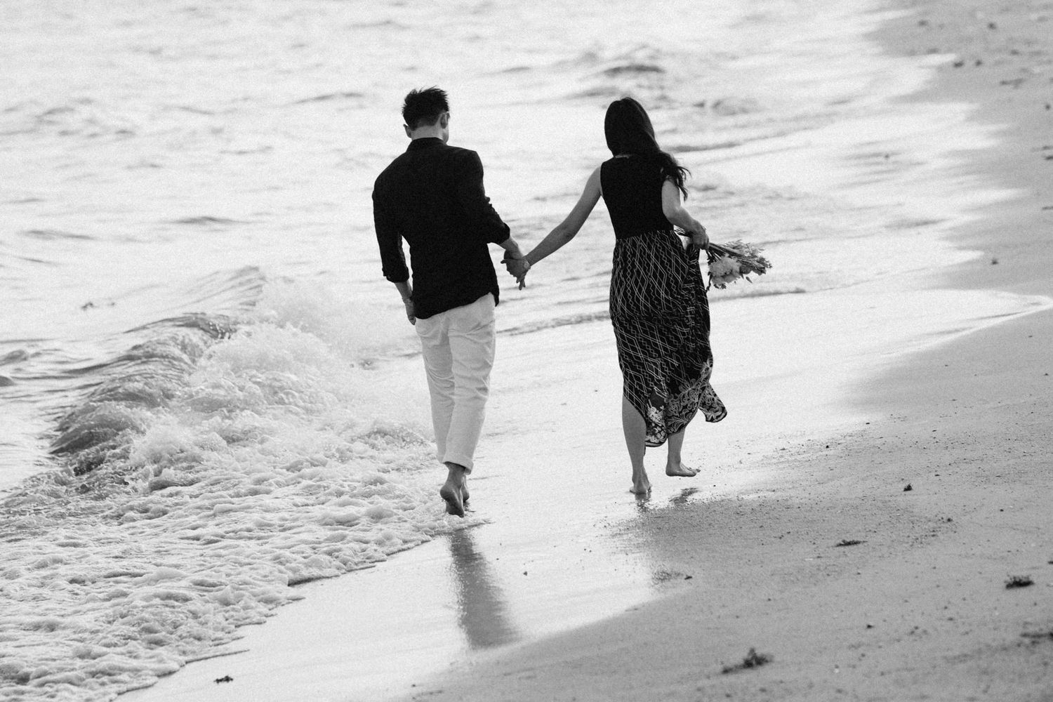 Black and white photo of couple strolling hand in hand on sandy beach.