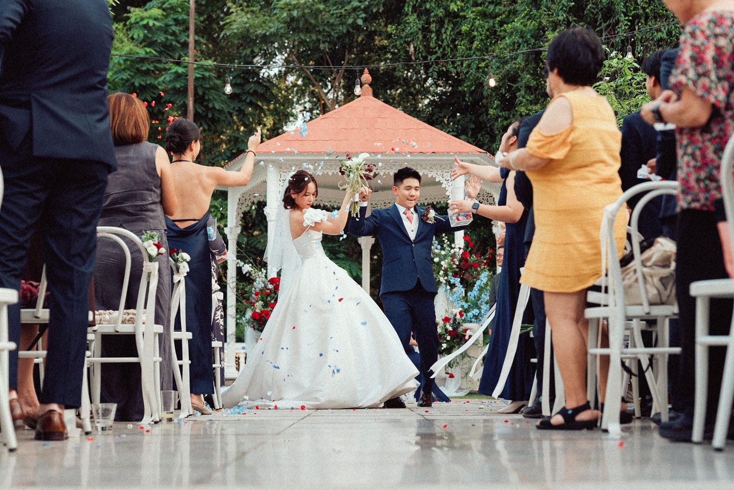 Wedding guests dance in a line formation outdoors during a festive celebration at a venue with white columns.