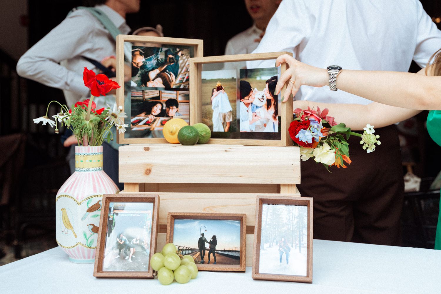 Wooden display boxes and frames showcasing family photos arranged on a table with flowers and fruit decorations.