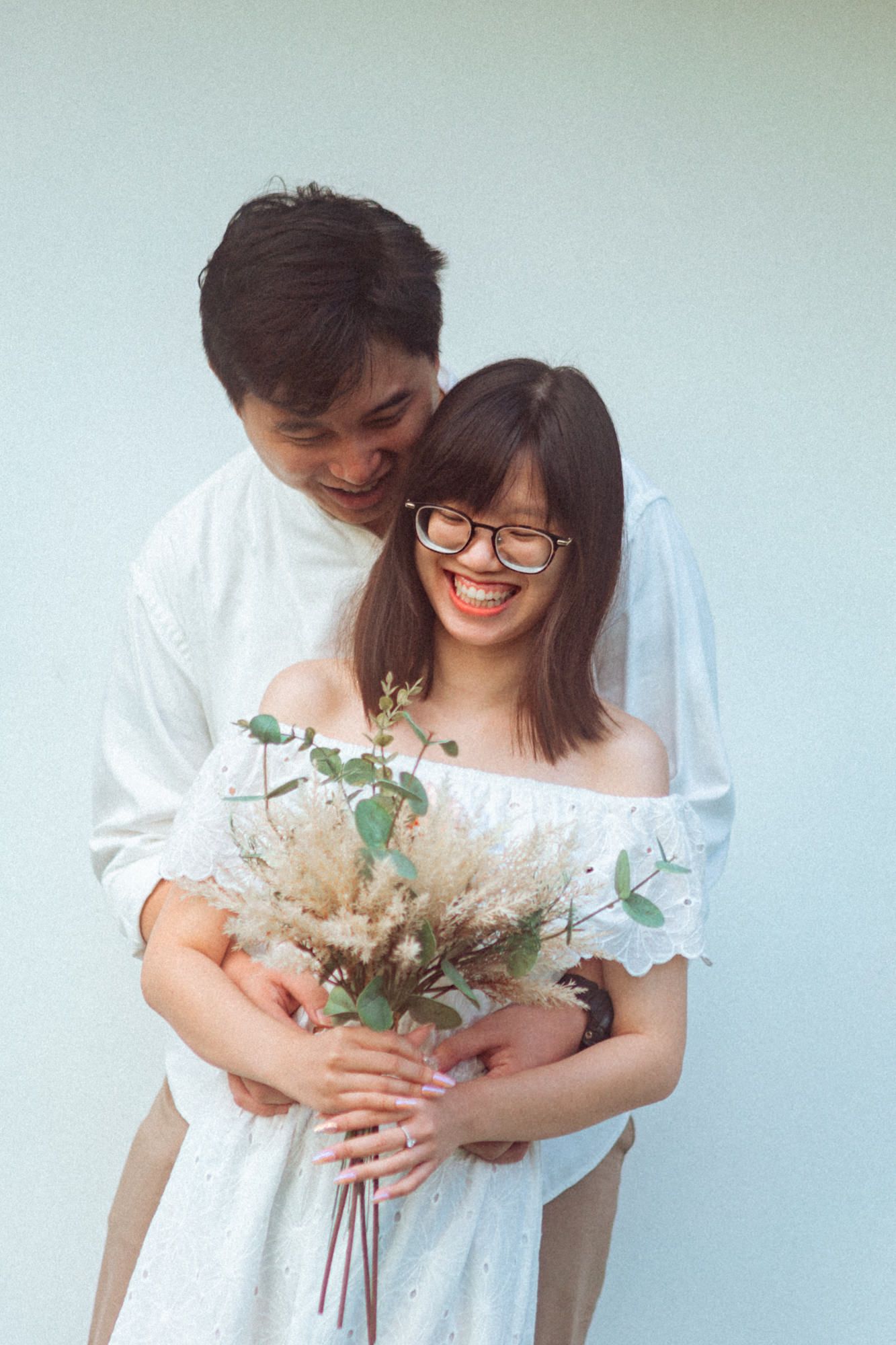 A couple embraces while holding a rustic bouquet of dried flowers against a light background.