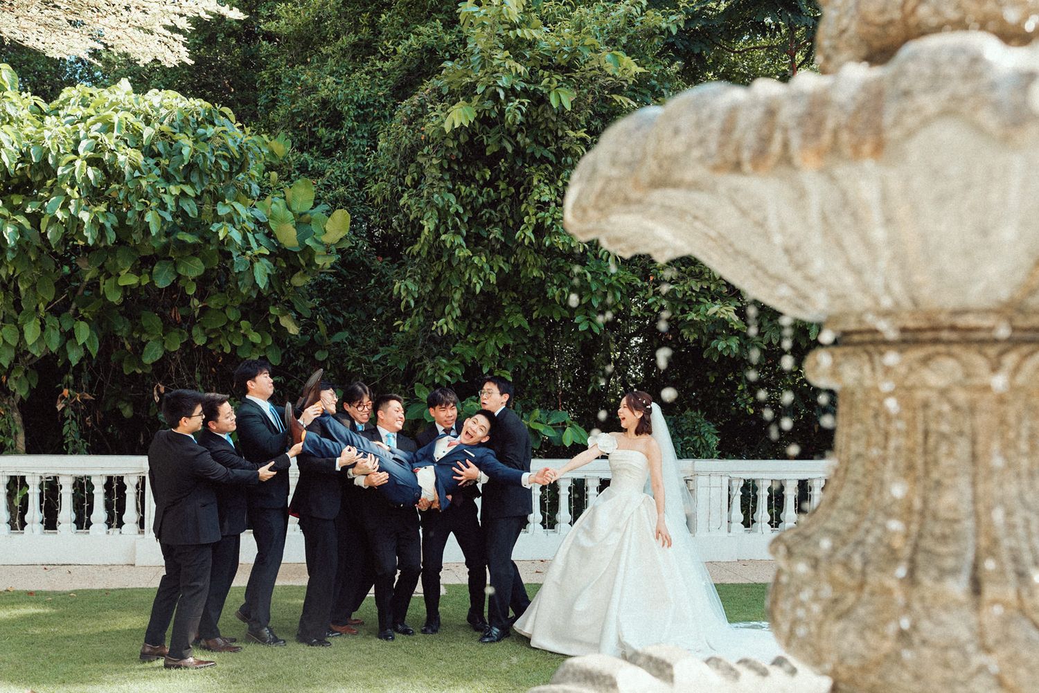 Wedding party poses playfully for photos near an ornate stone fountain in a garden setting.