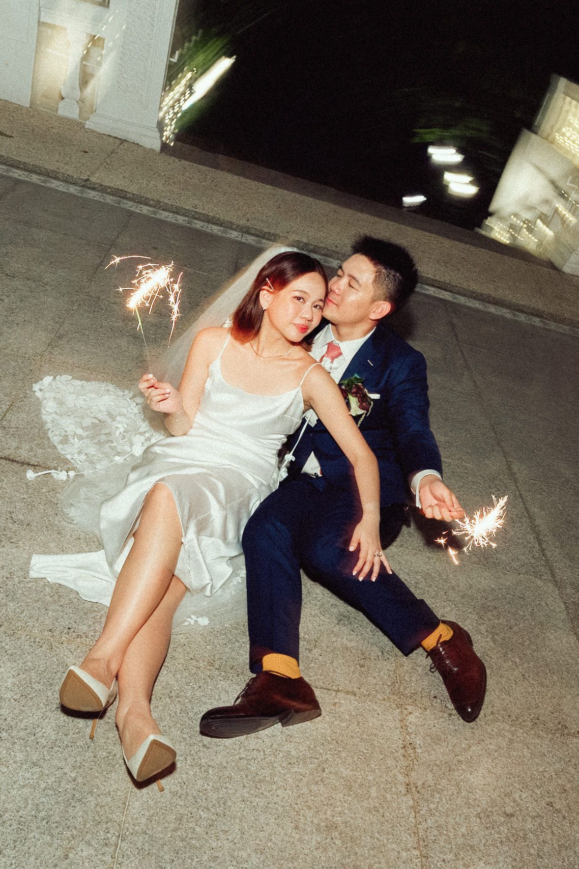 A couple in wedding attire sits on the ground holding sparklers during a nighttime celebration.