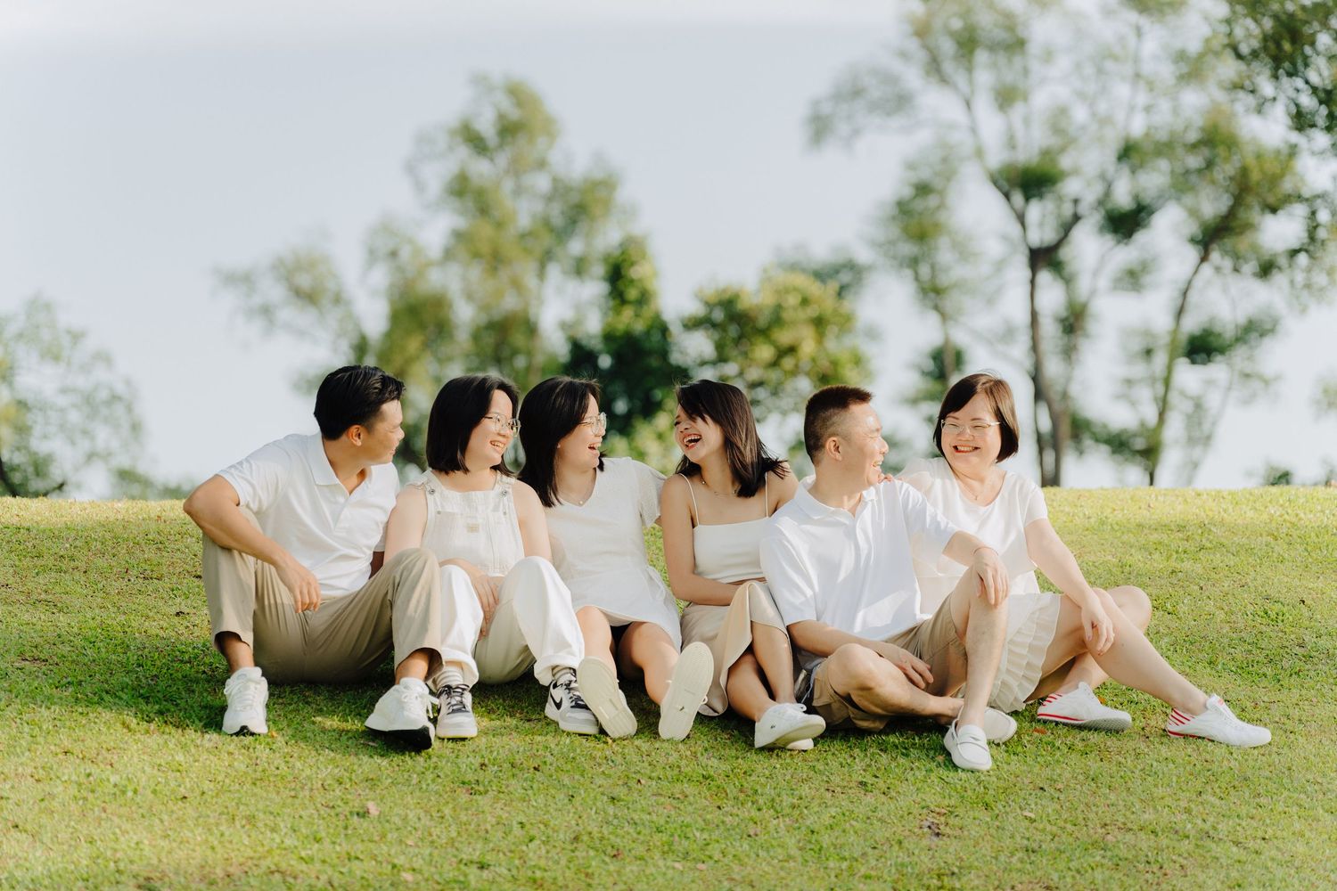 Group of friends sitting on grass in white outfits enjoying a sunny day outdoors.